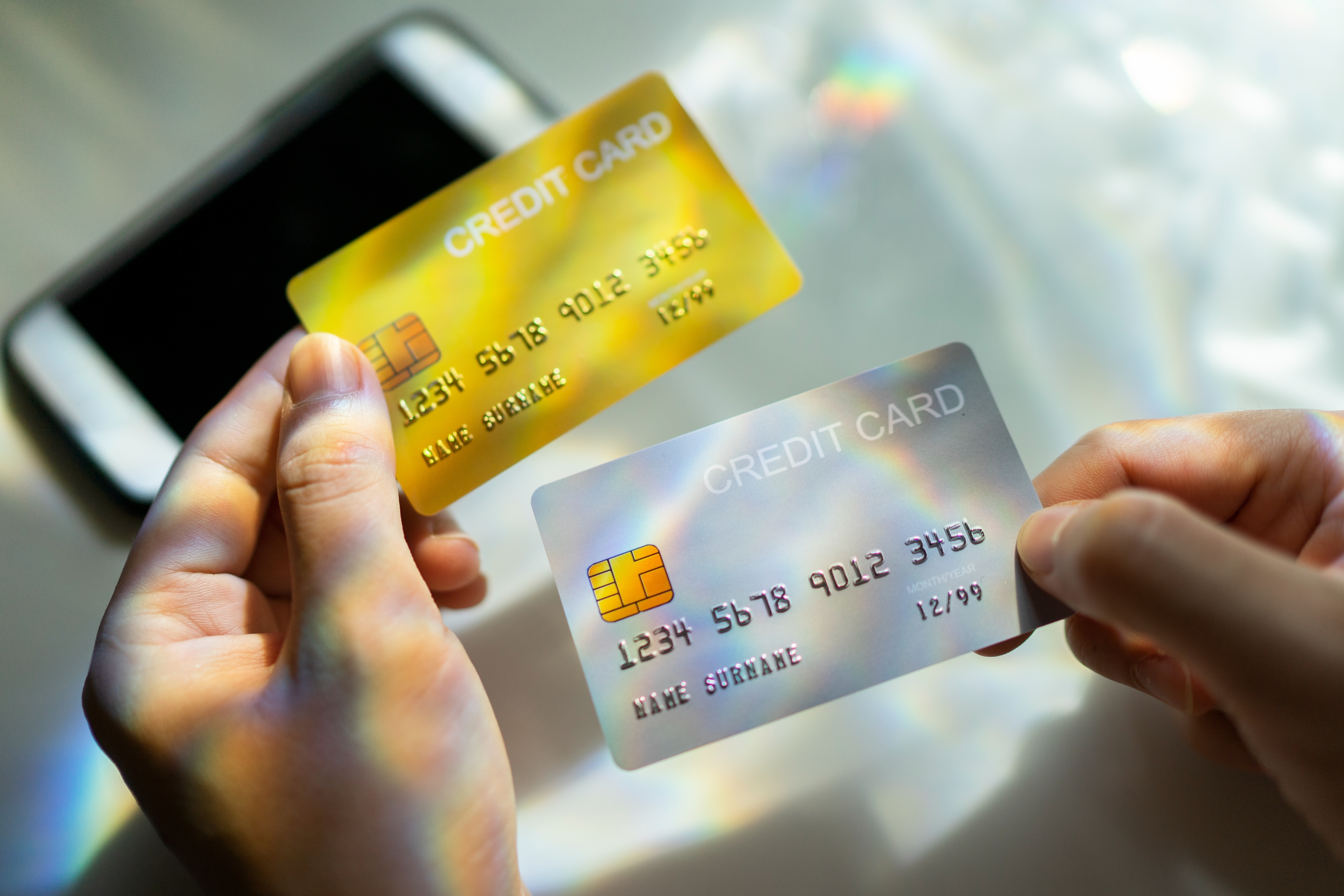 Two hands holding credit cards above a smartphone, with holographic light effects on the cards