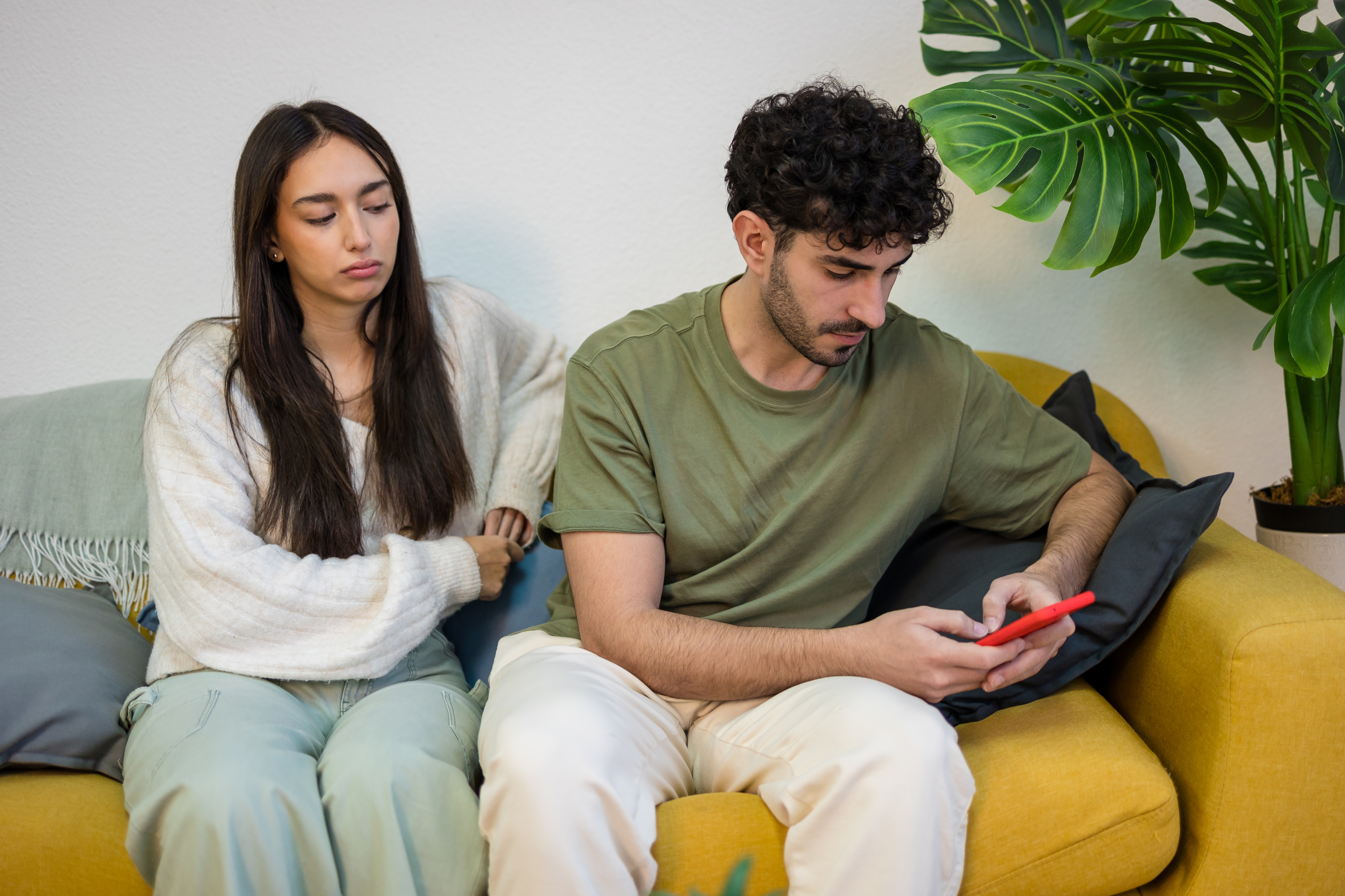A woman sits on a couch looking at a man on his phone, who is not paying attention to her