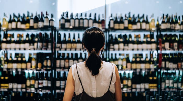 Person with a ponytail facing shelves filled with wine bottles in a store, choosing from a wide selection