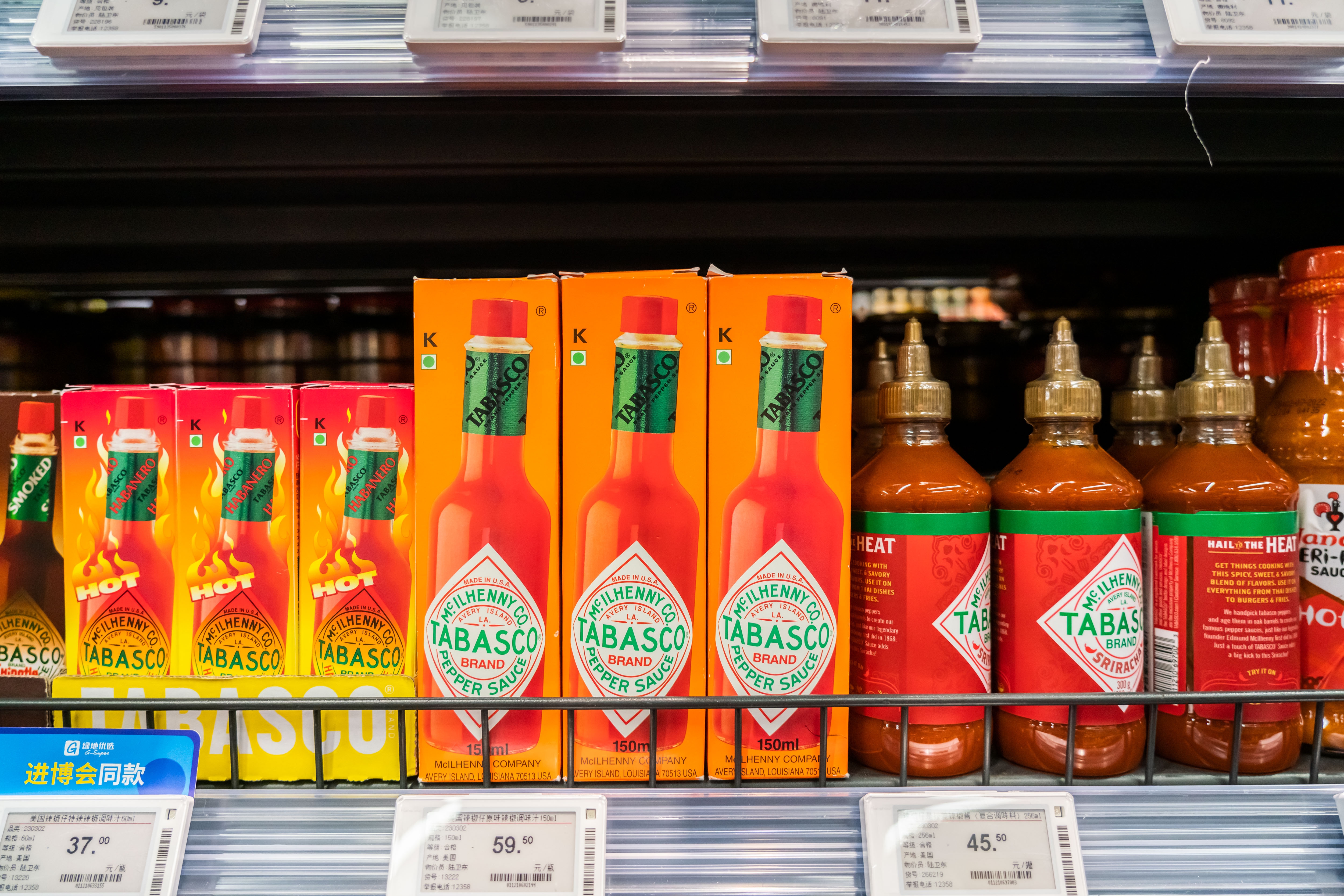 Bottles and boxes of Tabasco sauce displayed on a supermarket shelf. Price tags with various amounts are visible below the products