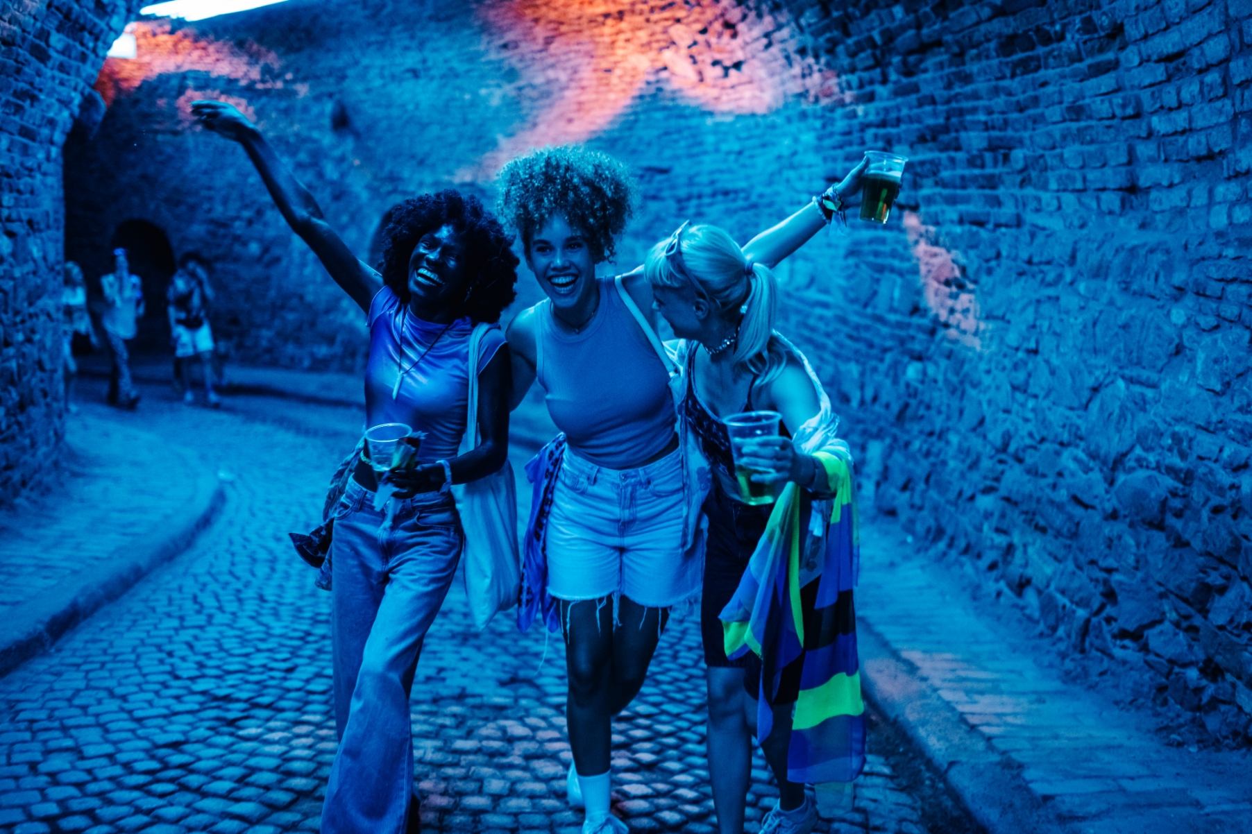 Three people joyfully walking in a cobblestone alley, each holding a drink, with one person draped in a colorful striped scarf