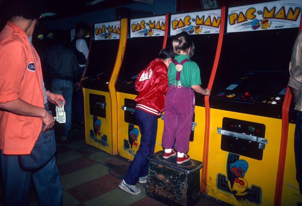 Two children playing Pac-Man at a vintage arcade, one wearing a red jacket and the other in purple overalls standing on a step stool