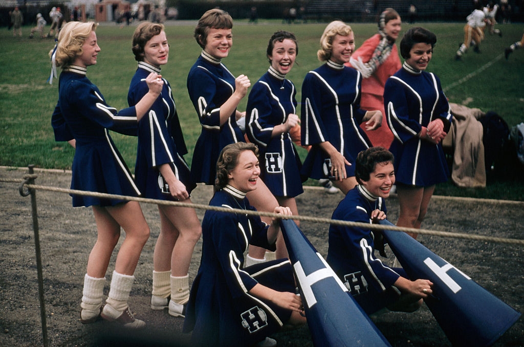 Retro cheerleaders in matching blue outfits with letter "H" enjoy a moment of camaraderie on a sports field, holding megaphones