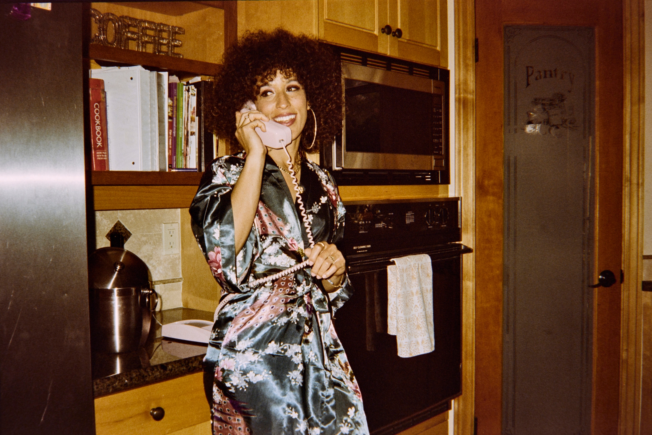 Person in floral robe smiles while talking on a retro phone in a kitchen