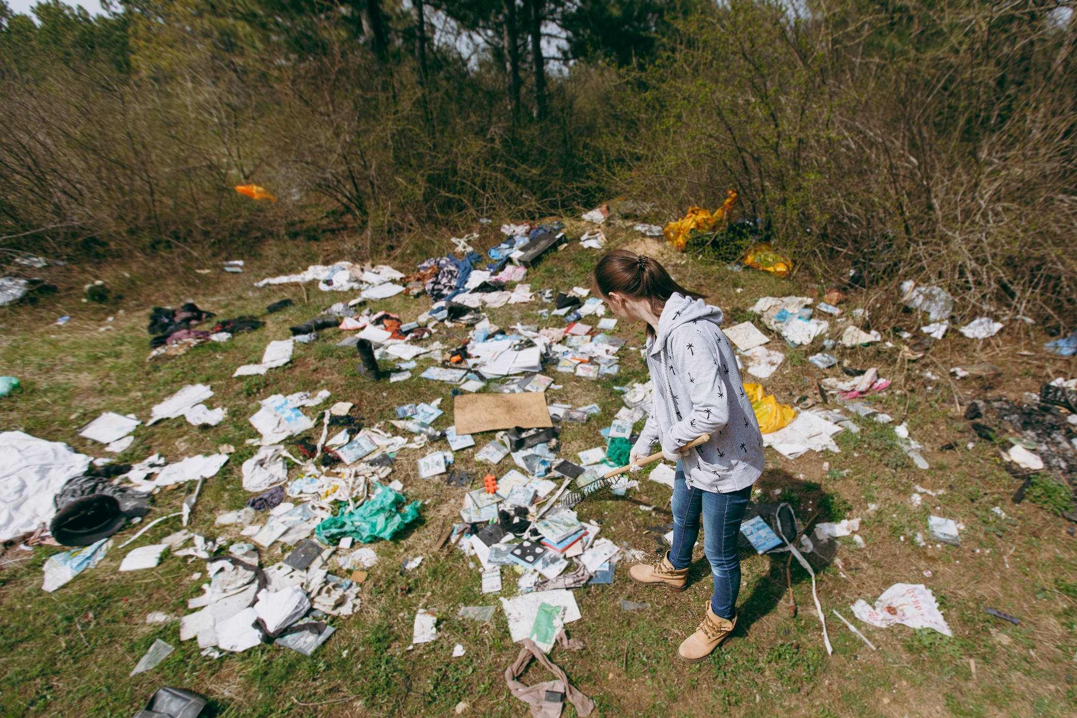Person standing in a field littered with trash, surrounded by trees, examining the scattered debris