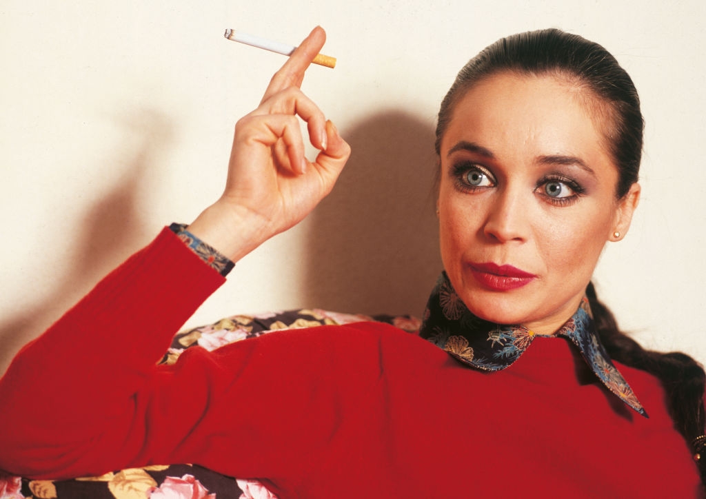 Person posing with a cigarette holder, wearing a floral-collared shirt and red top, staring intently