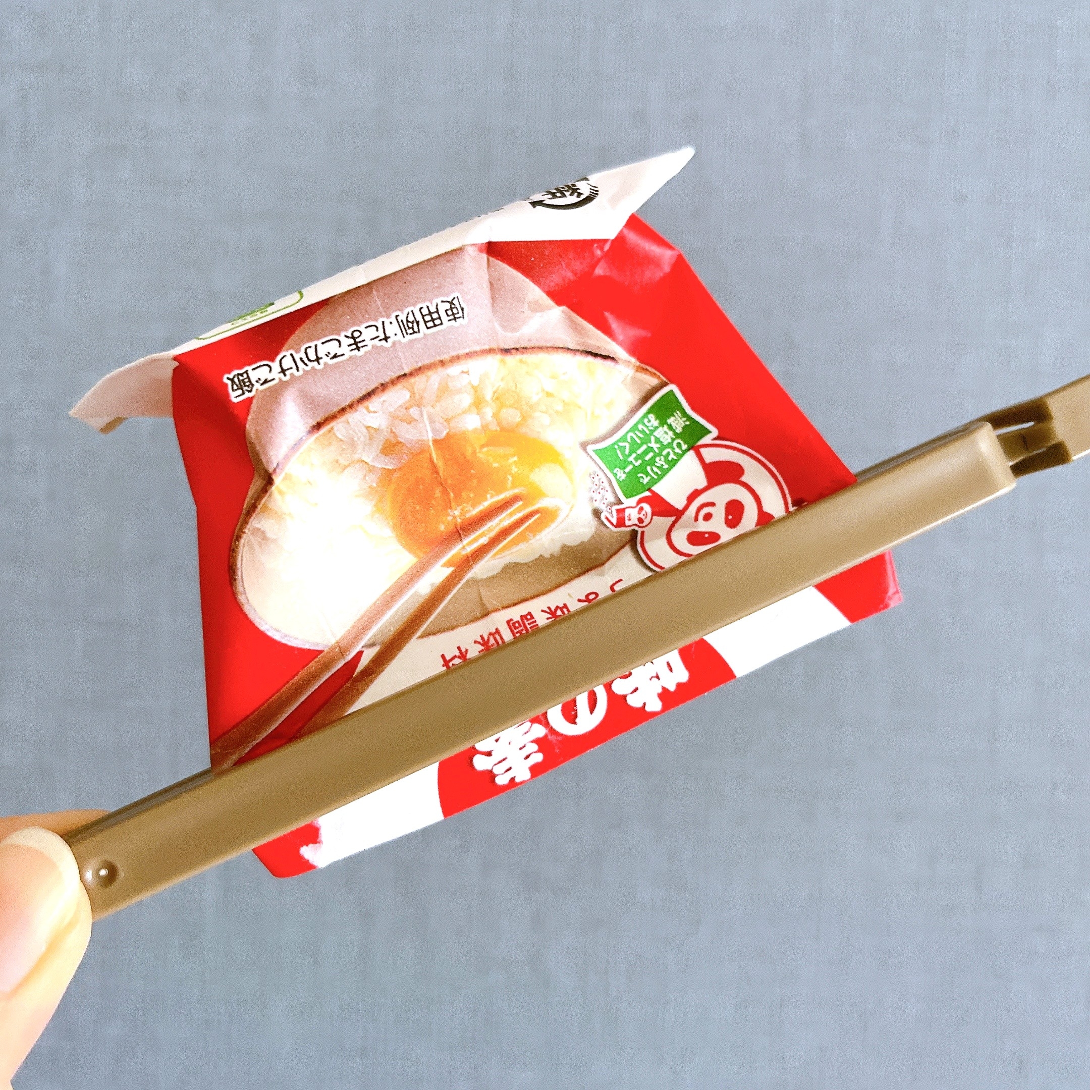 白い袋入りの赤い容器に半分割れたプリンのイラスト。箸で持ち上げるためのホルダーが手に持たれている。