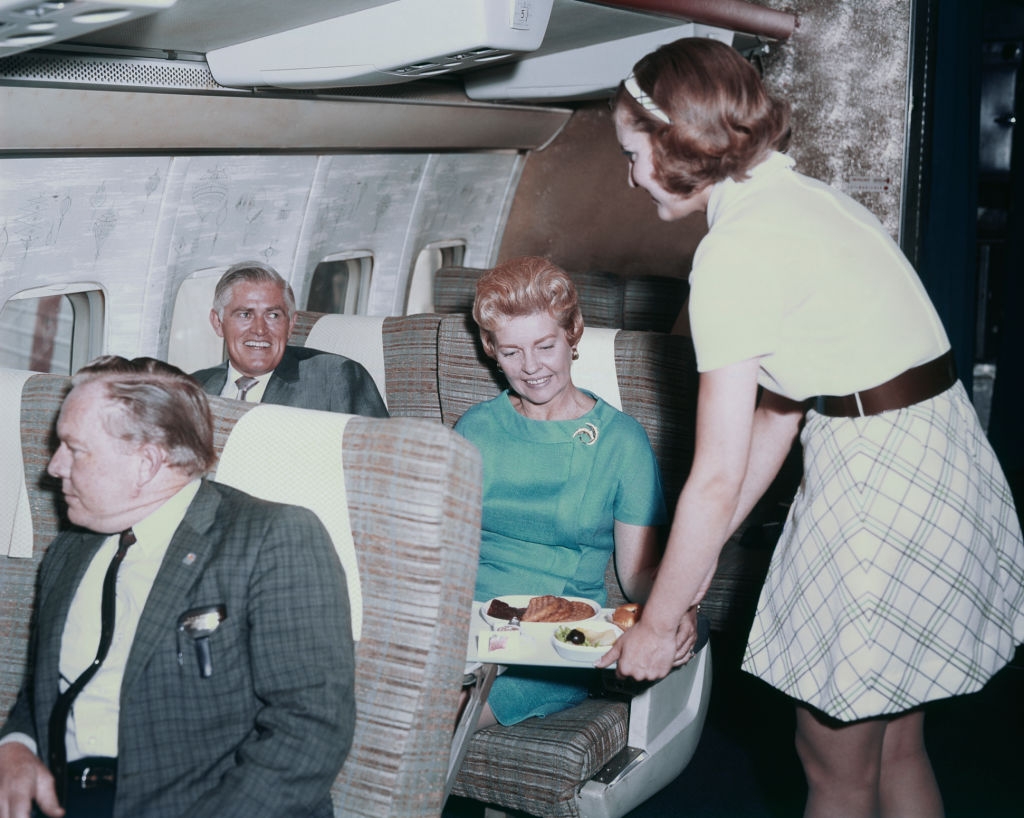 Passengers seated in a vintage airplane cabin are served by a flight attendant. They appear relaxed and engaged in conversation