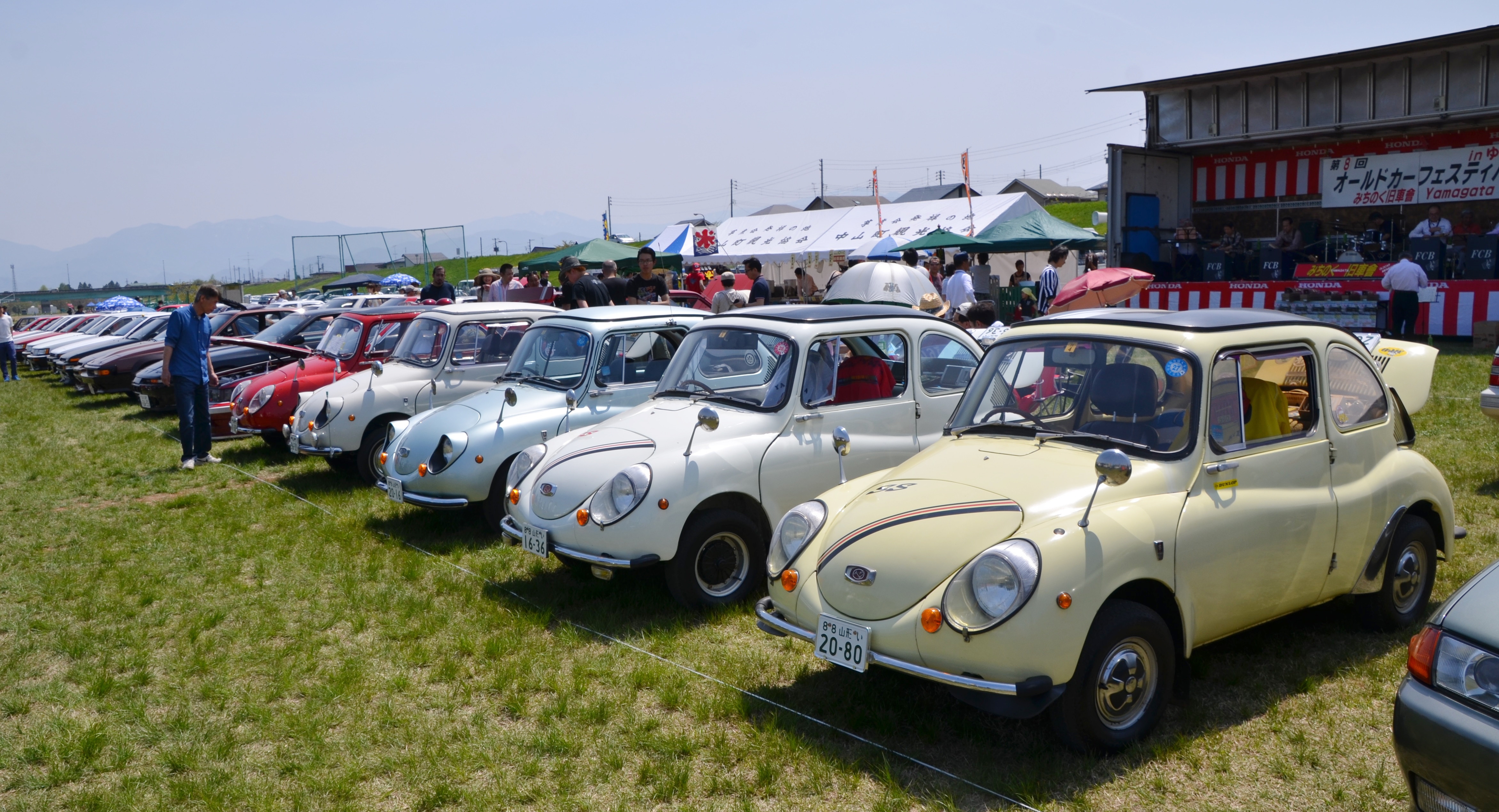 整然と並ぶクラシックカーの展示会。観客が見学し、販売ブースも見える。