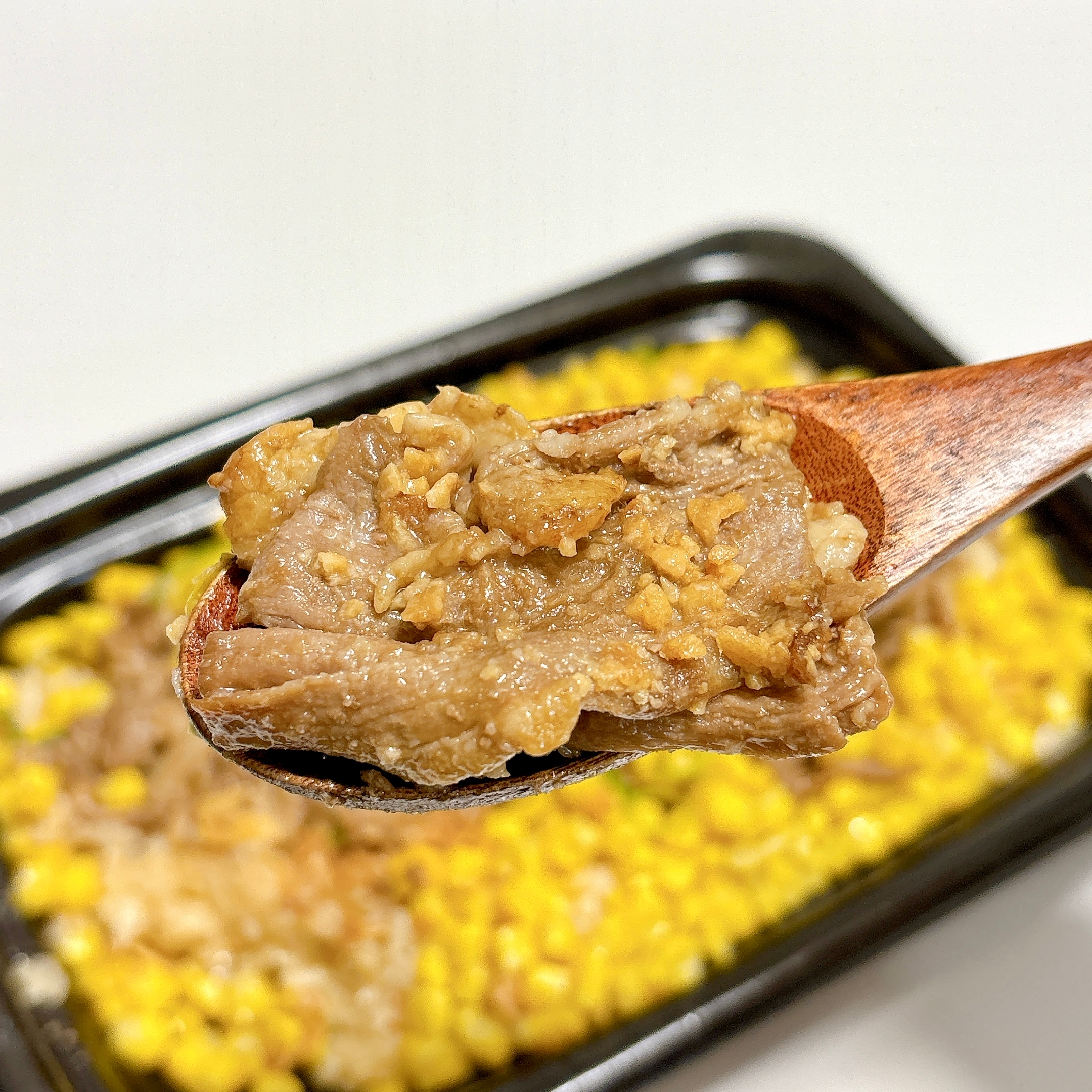 スプーンにのった肉とコーン炒めのクローズアップ。背景には黒いトレーに盛り付けられたコーンが見える。