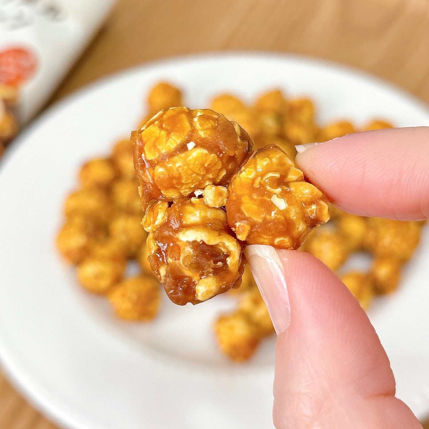 キャラメルポップコーンを指でつかんでいる手。背景にお皿の上のポップコーンが写っている。