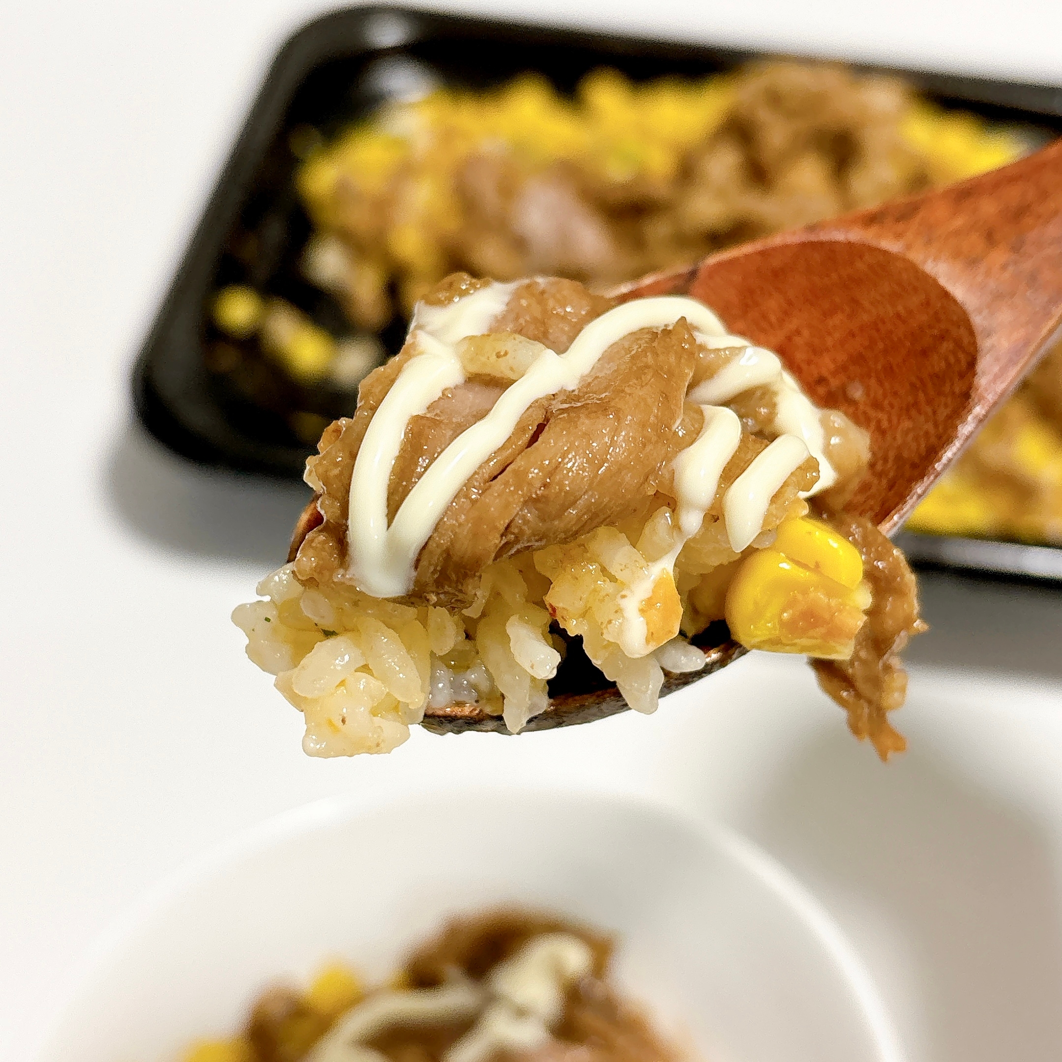 スプーンに乗ったご飯、コーン、マヨネーズ、焼肉がアップで映っている。背景にトレイに載った同じ料理が見える。