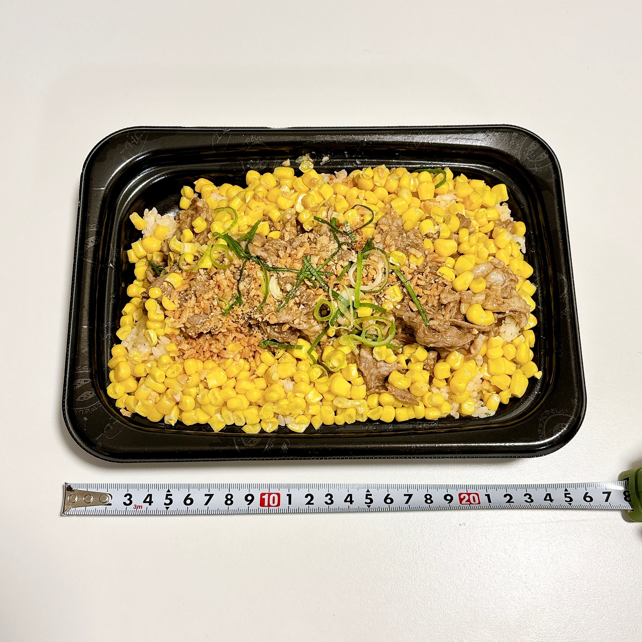 お弁当容器に盛られたトウモロコシと肉の料理、上に刻んだネギが乗せられています。横に定規が置かれています。