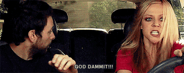 A woman, frustrated, yells while driving a car with a man in the passenger seat. Subtitles read "GOD DAMMIT!!!"