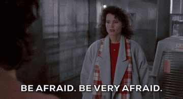 Woman wearing a coat and scarf appears frightened as she stands in a dimly lit setting with text overlay: "Be afraid. Be very afraid."