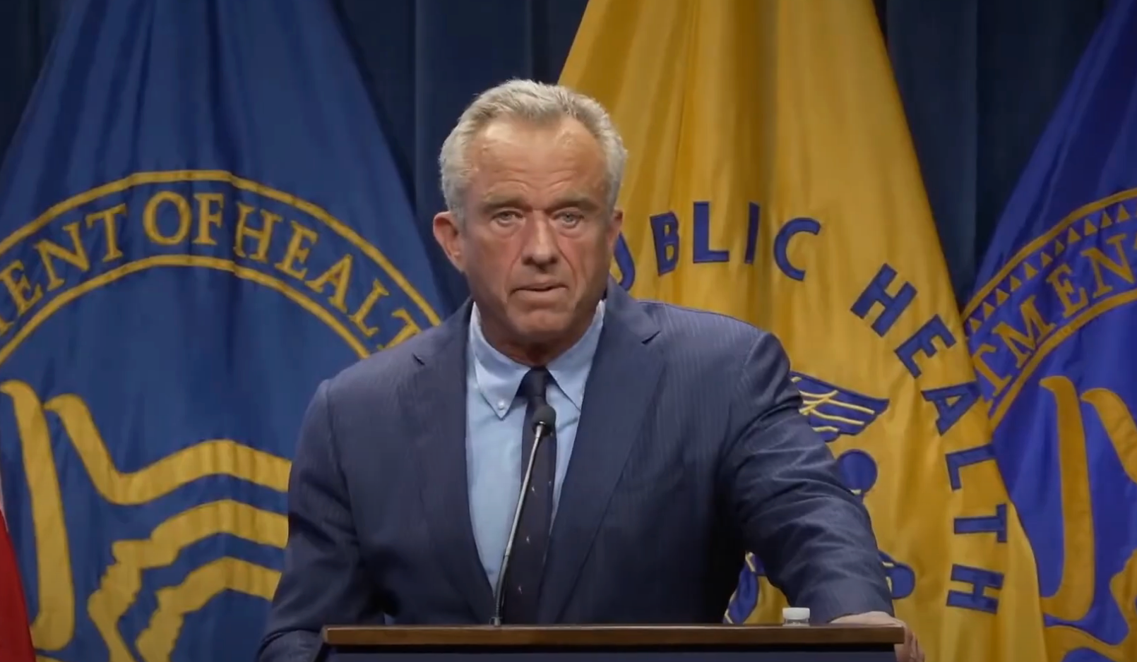 A man in a suit speaks at a podium with public health department seals in the background
