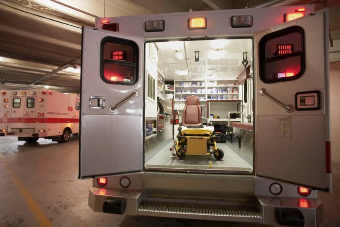 The image shows the open back of an ambulance parked indoors, with a view of its interior and a stretcher inside. Another ambulance is in the background