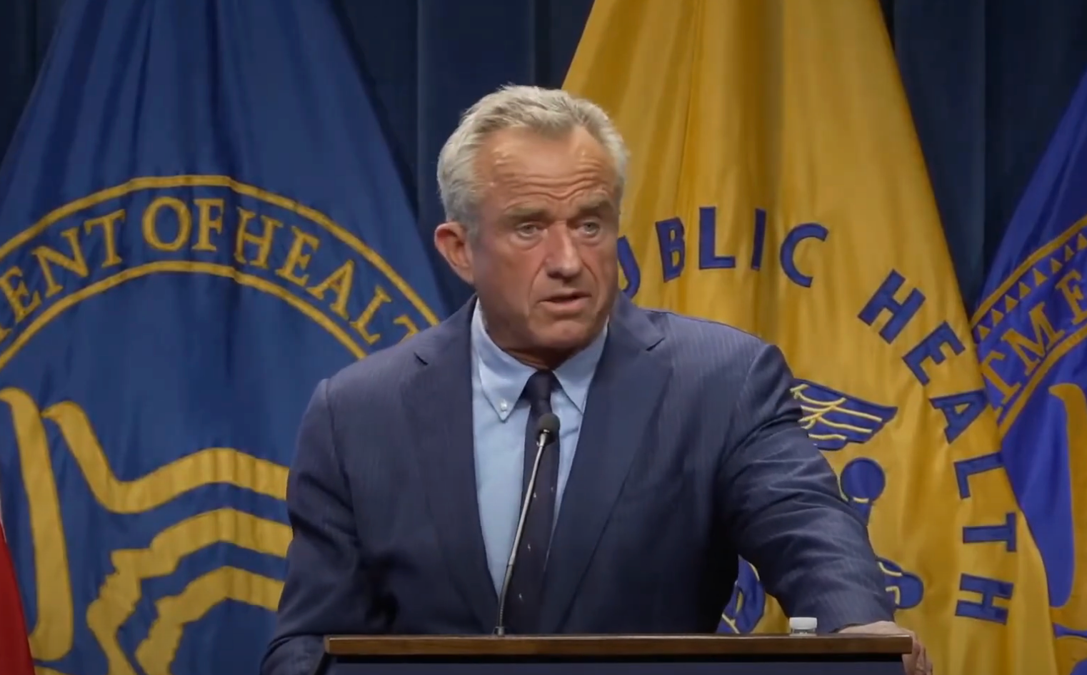A man in a suit speaks at a podium, in front of public health-themed banners