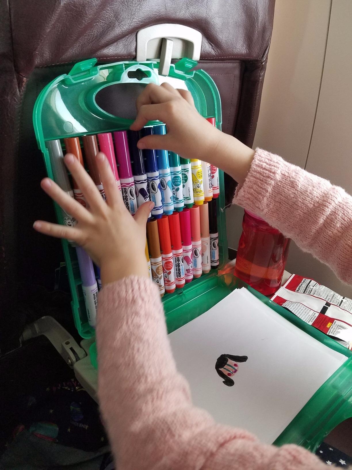 Child's hands organizing markers in a case on a plane tray table, next to a water bottle and bag
