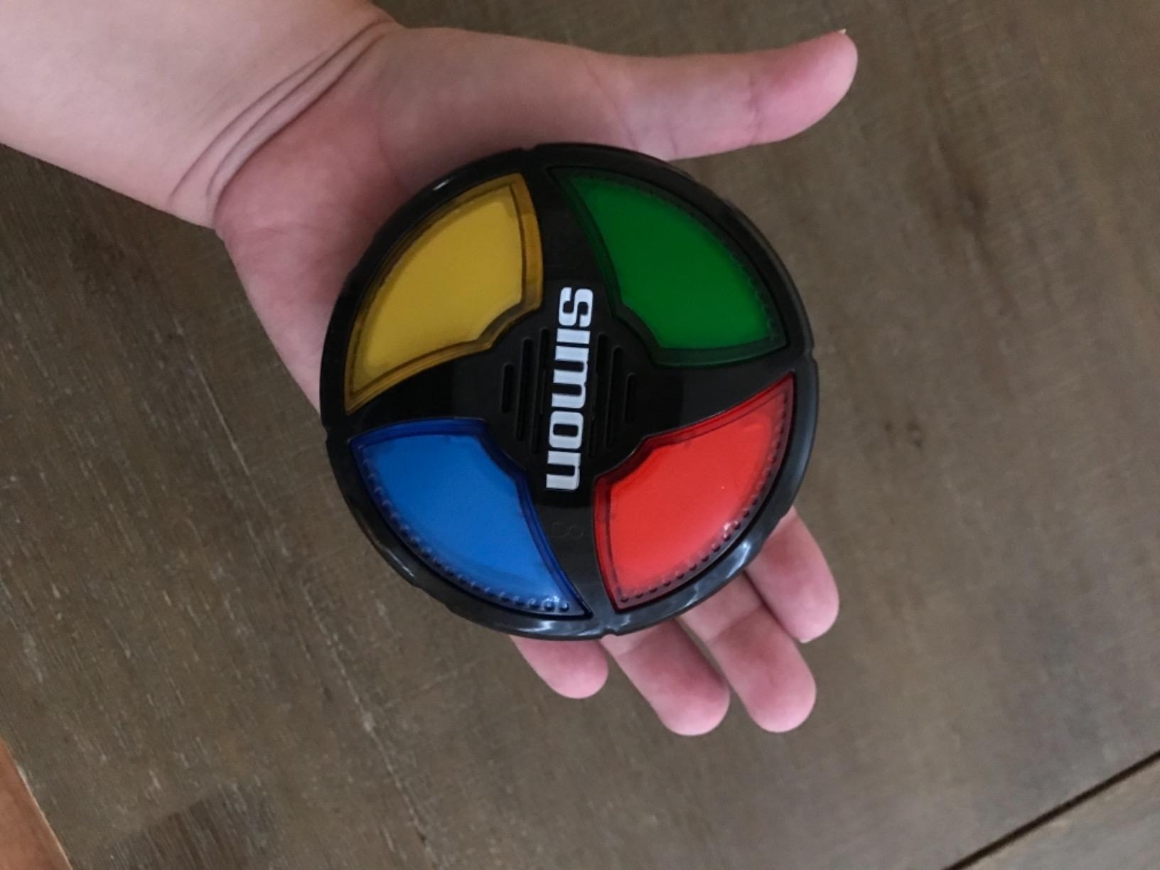 Hand holding Simon game, a round electronic memory game with red, green, blue, and yellow buttons