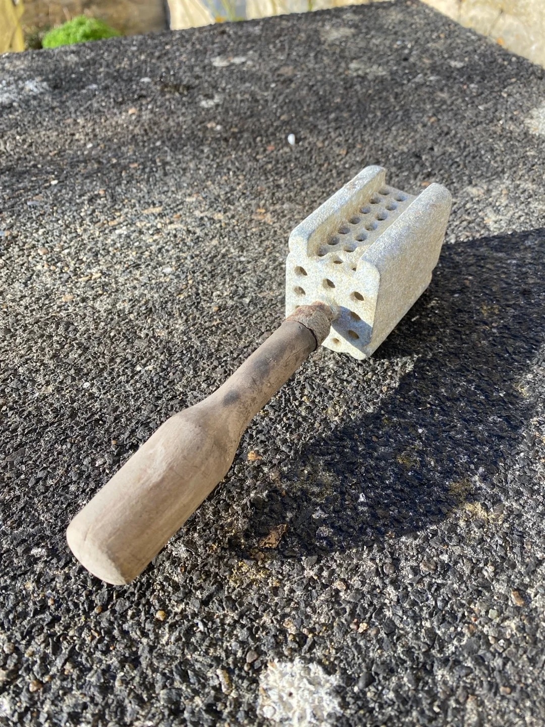Hammer with a wooden handle stuck into a brick with a hole pattern, lying on a rough surface