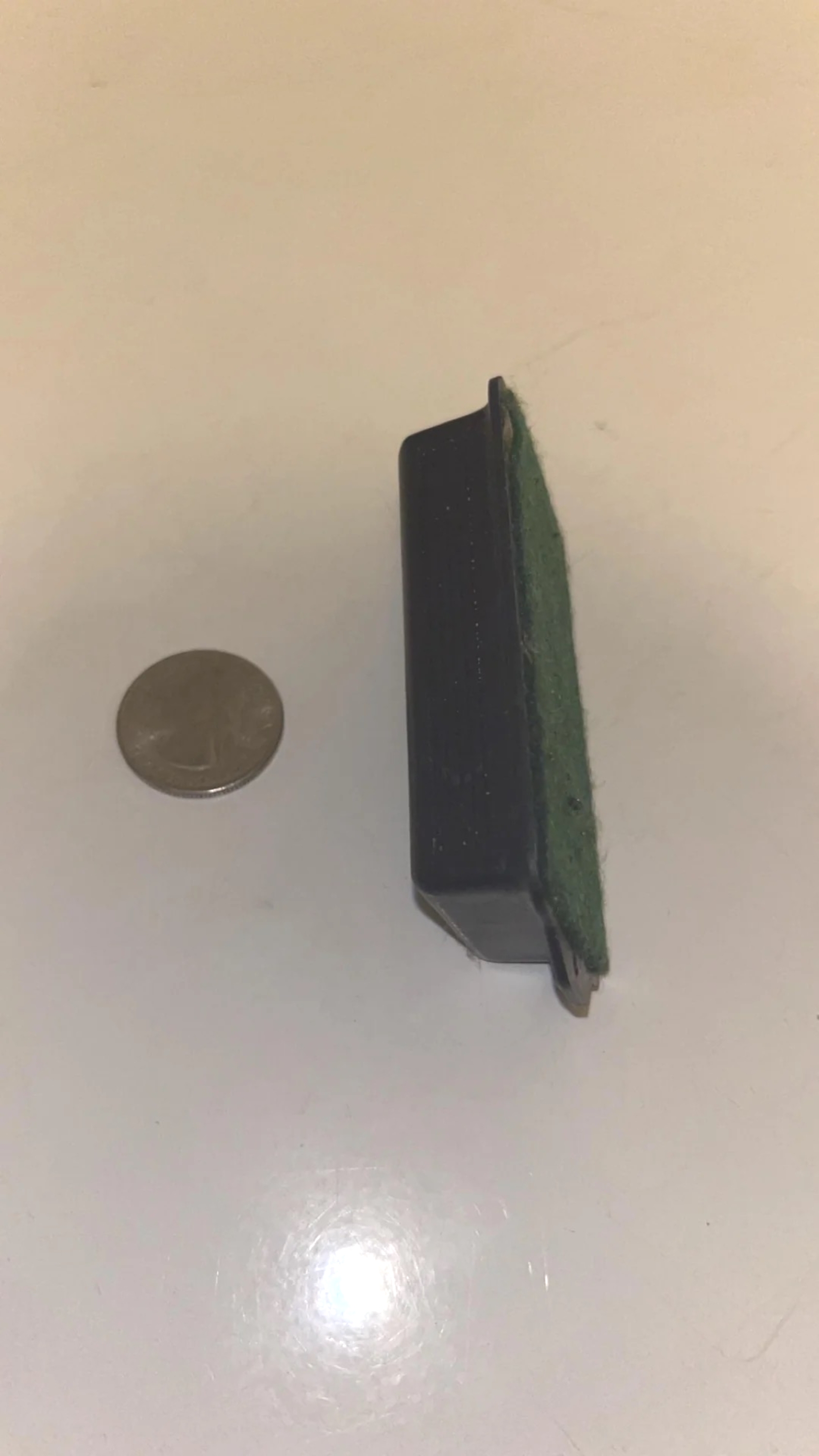 A small rectangular object with a green felt side sits next to a coin for scale on a light surface