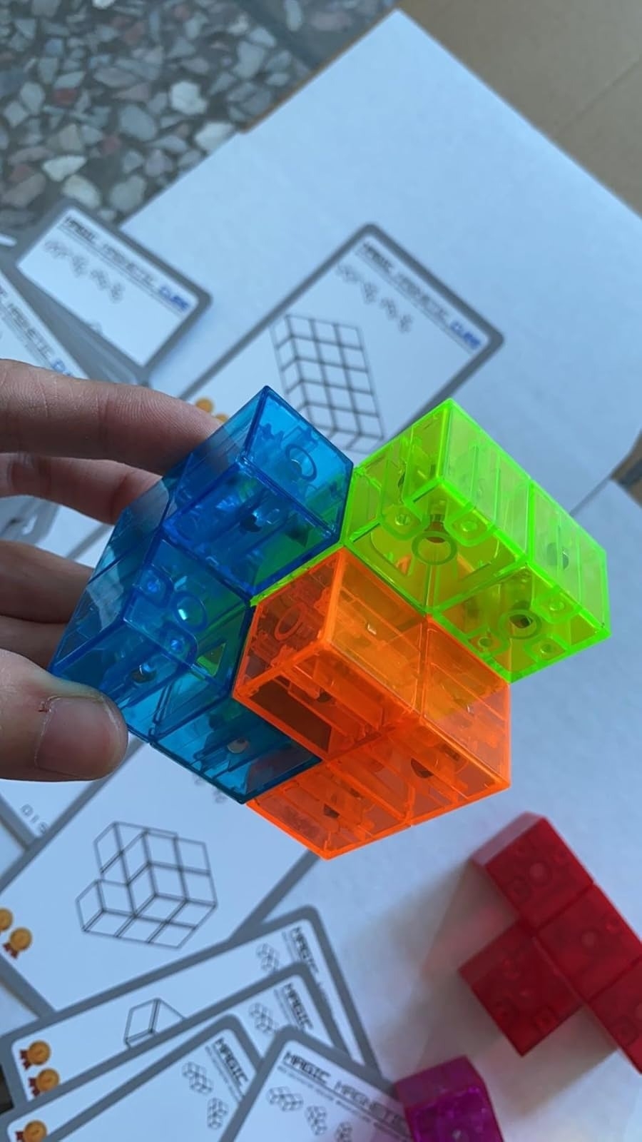 Hand holds colorful transparent cube puzzle pieces, with instruction sheets in the background on a table
