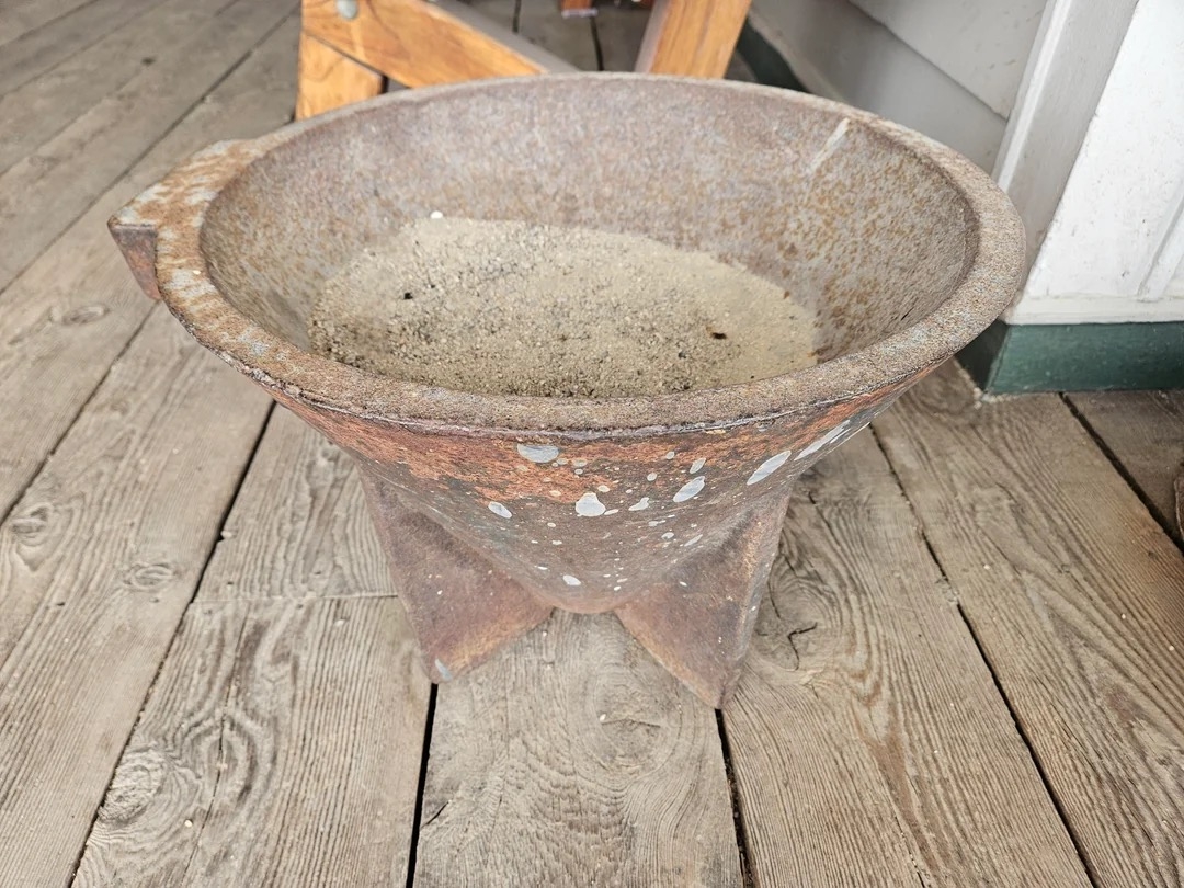 A rustic, weathered metal bowl sits on a wooden deck. It's filled with sand and shows signs of age and use