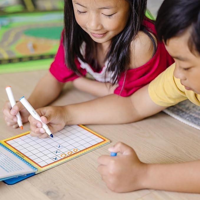 Two children lying on the floor, happily playing tic-tac-toe with colored markers on a grid notebook