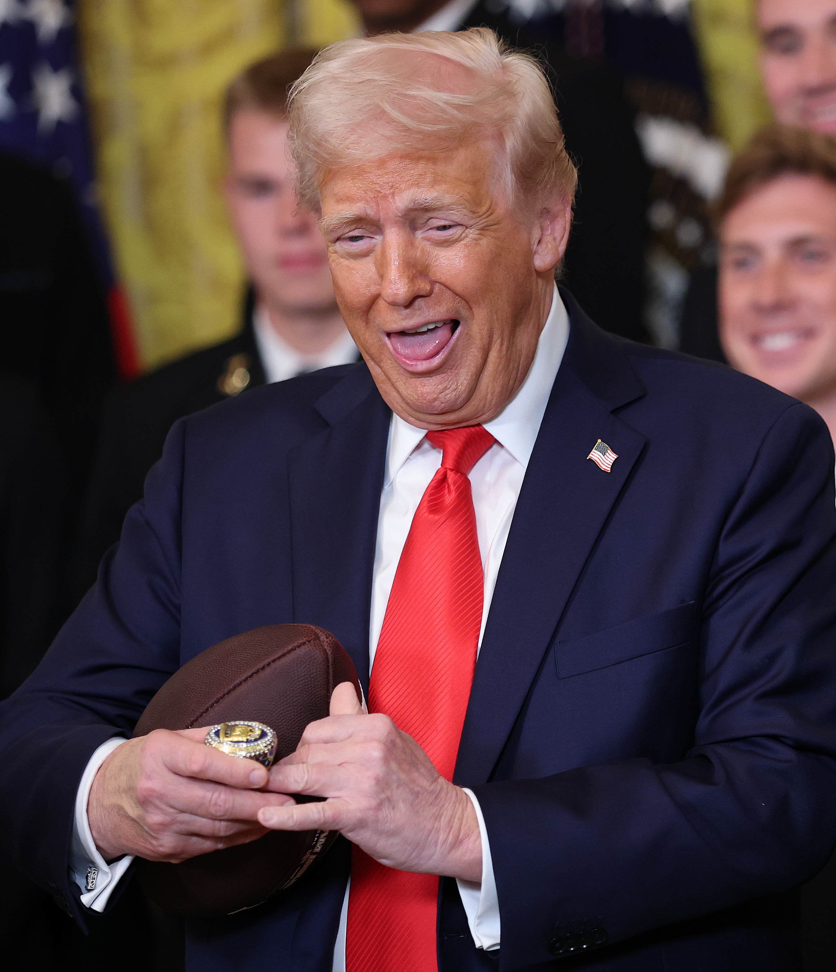 Person in a suit holding a football and a ring, smiling, surrounded by people in formal attire at an event