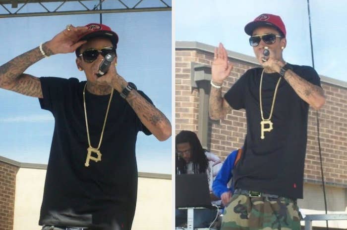 Person performing on an outdoor stage, wearing a black t-shirt, red cap, sunglasses, large pendant necklace, and camo pants, holding a microphone