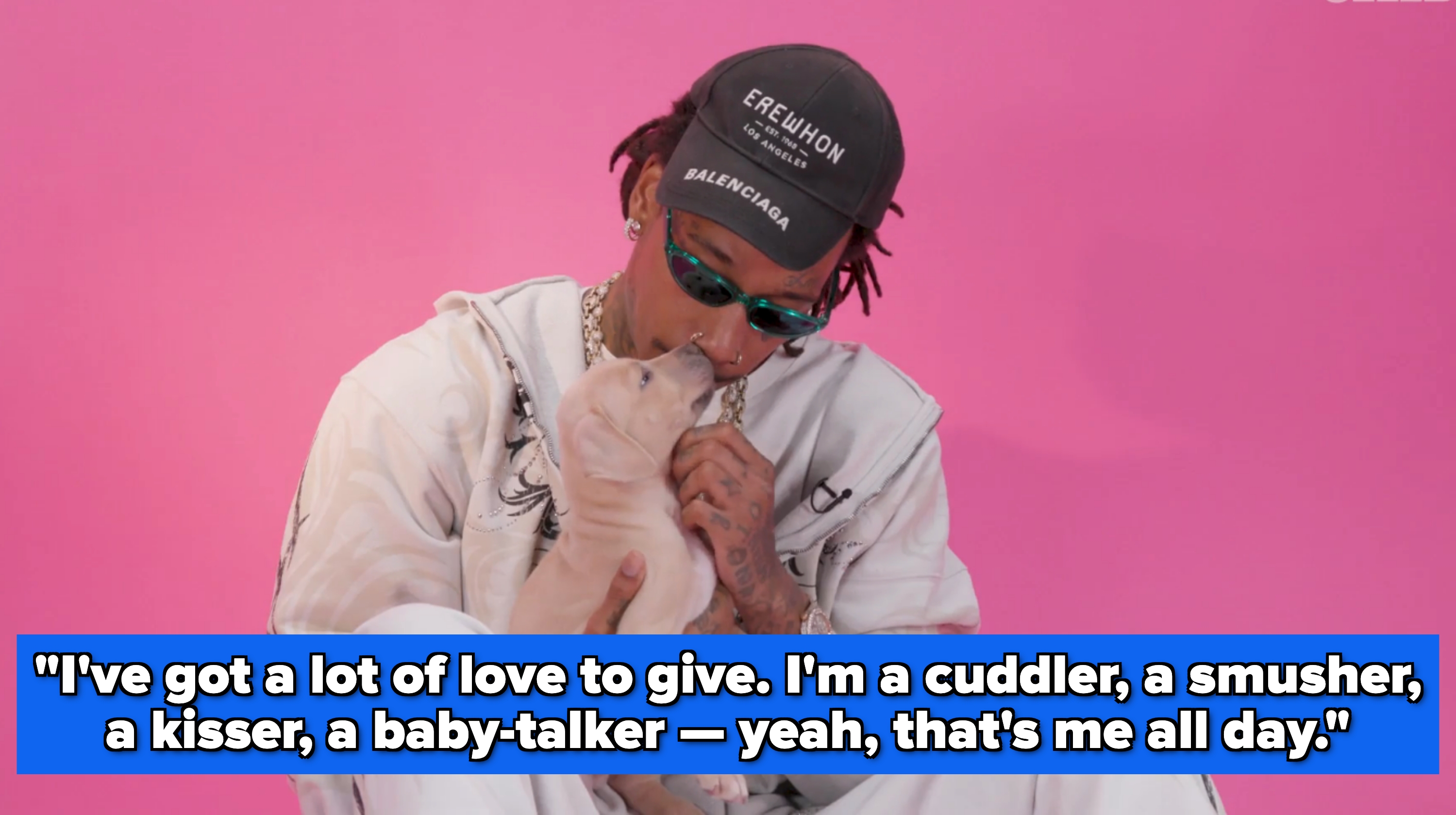 A person in a casual tracksuit and cap kisses a puppy against a pink background
