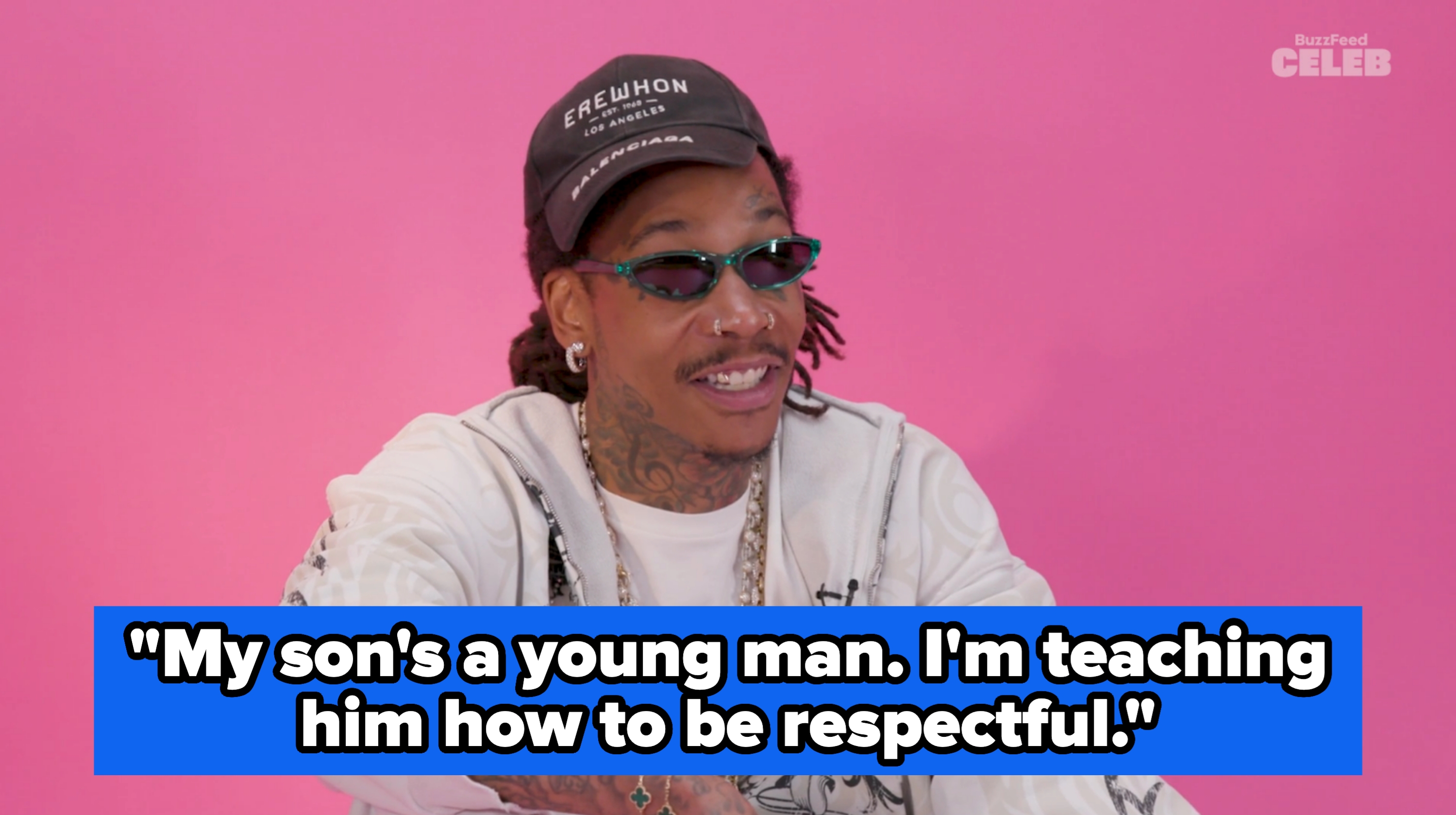 Person seated against a plain background wearing a hat, sunglasses, patterned jacket, and layered chain necklaces, smiling during an interview