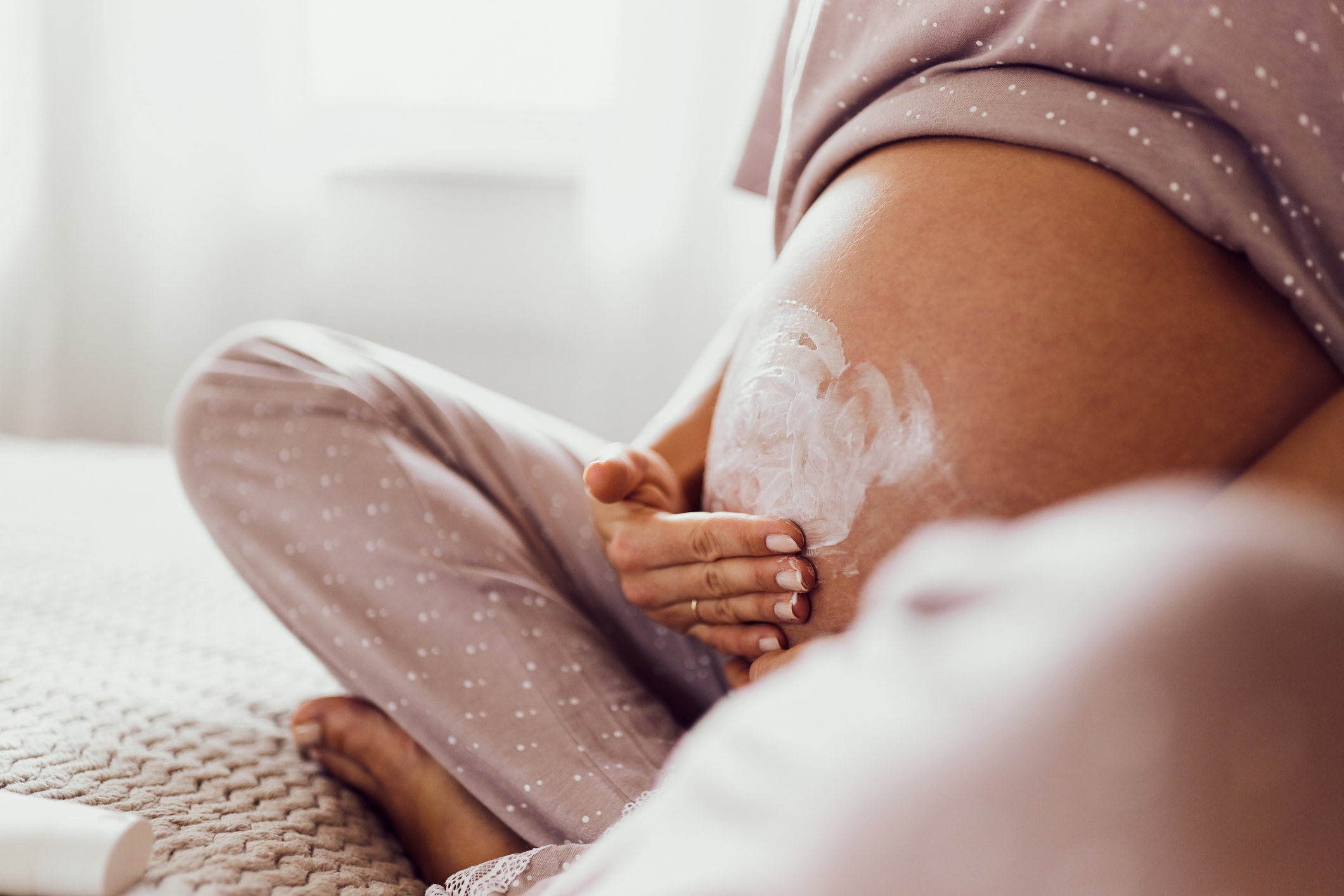 A person in comfortable home attire applies lotion to their pregnant belly while sitting on a cozy bed
