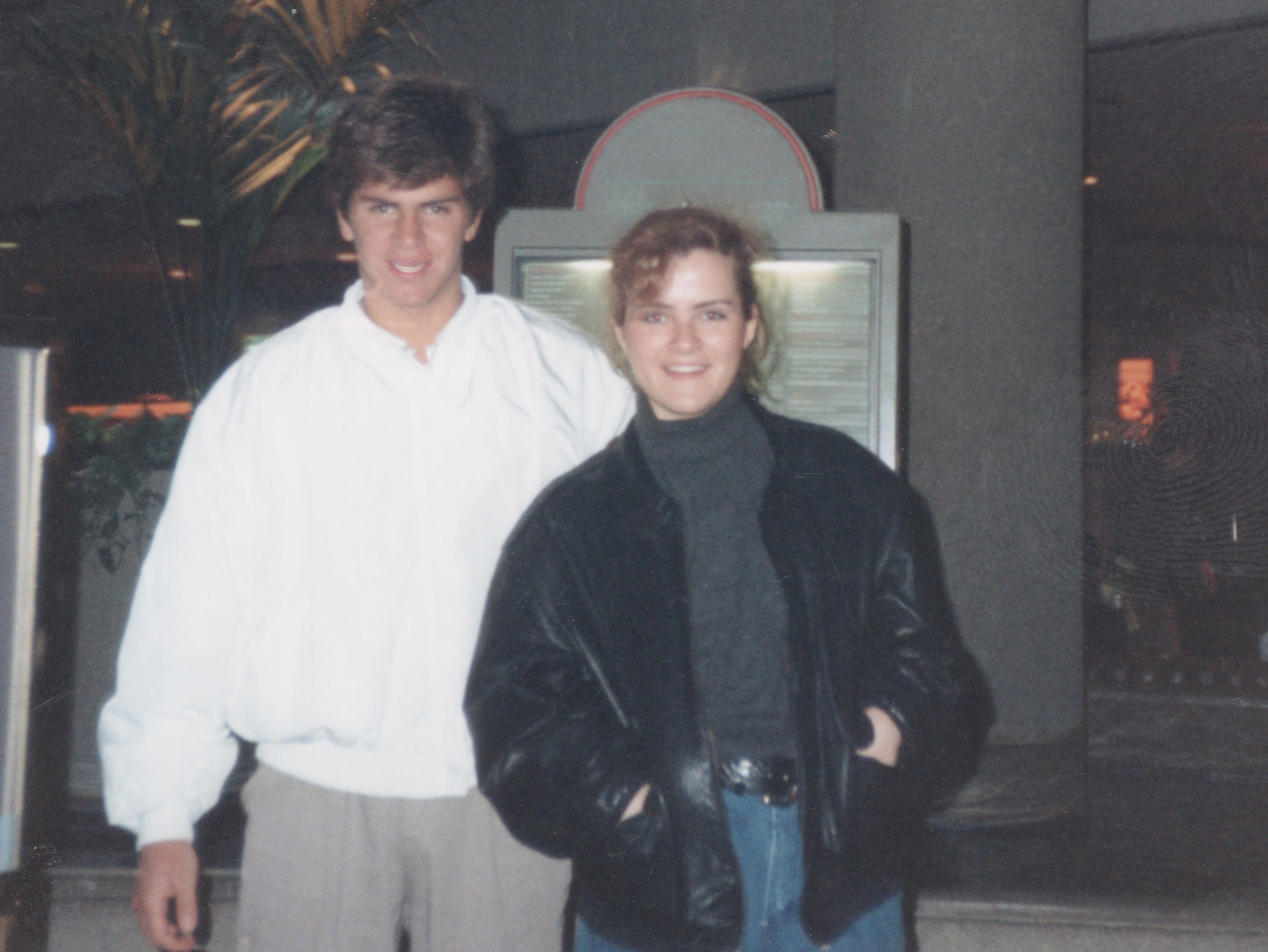 Two people smiling indoors, one wearing a white shirt, the other in a black jacket and turtleneck