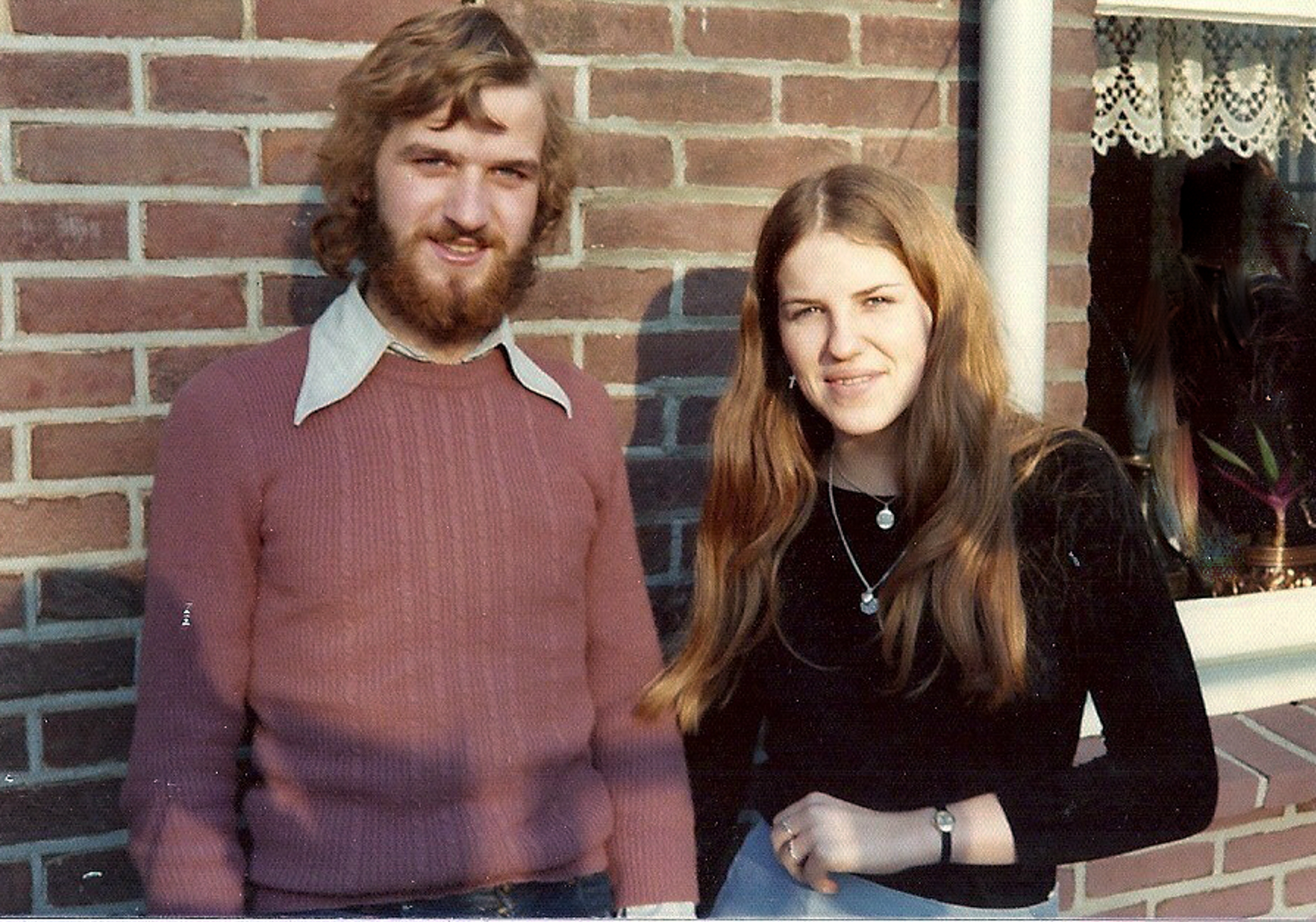 A couple standing together outside by a brick wall, both smiling. The man has a beard and the woman has long hair, wearing casual clothing