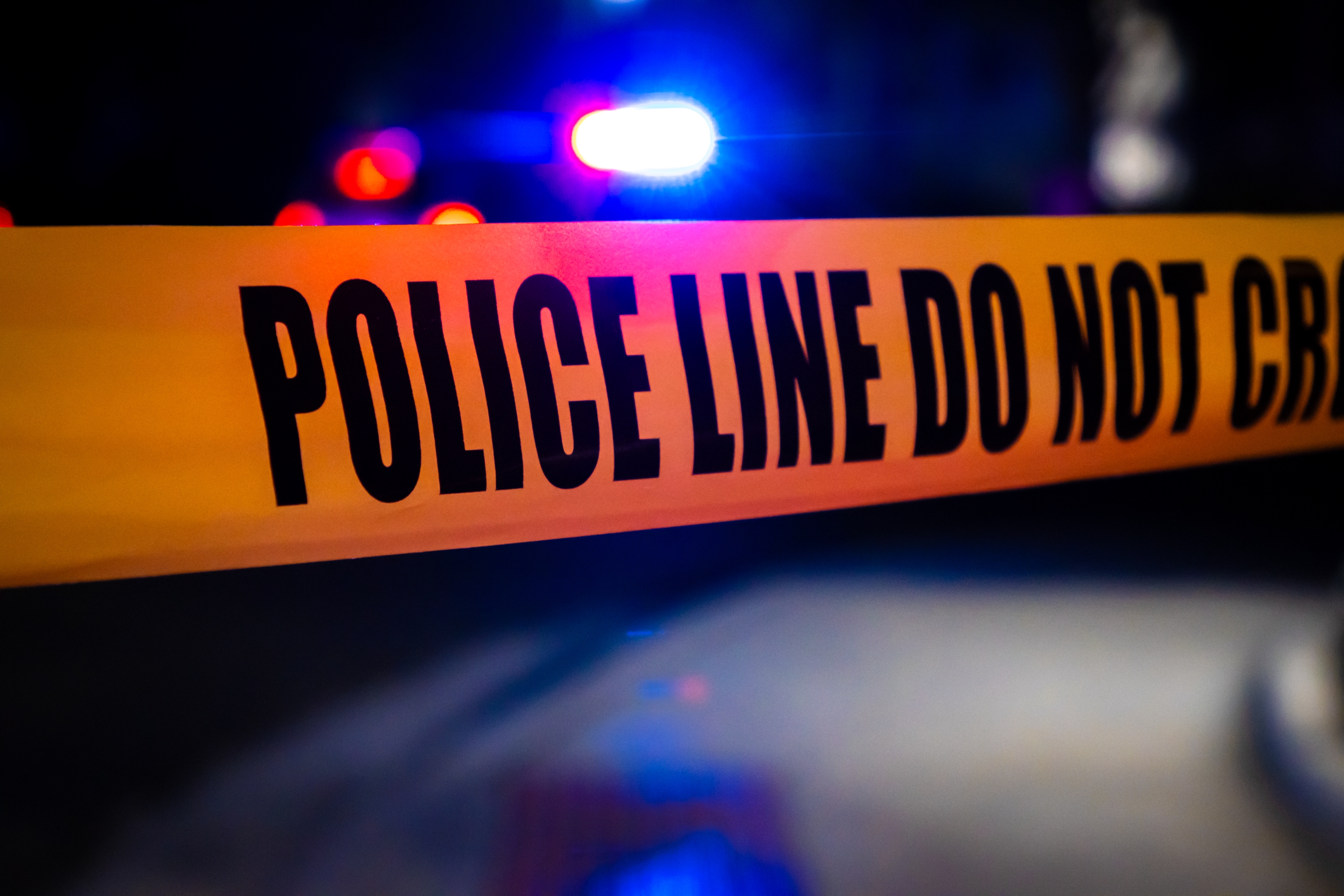 Close-up of police tape with &quot;POLICE LINE DO NOT CROSS&quot; text visible; blurred emergency lights in the background