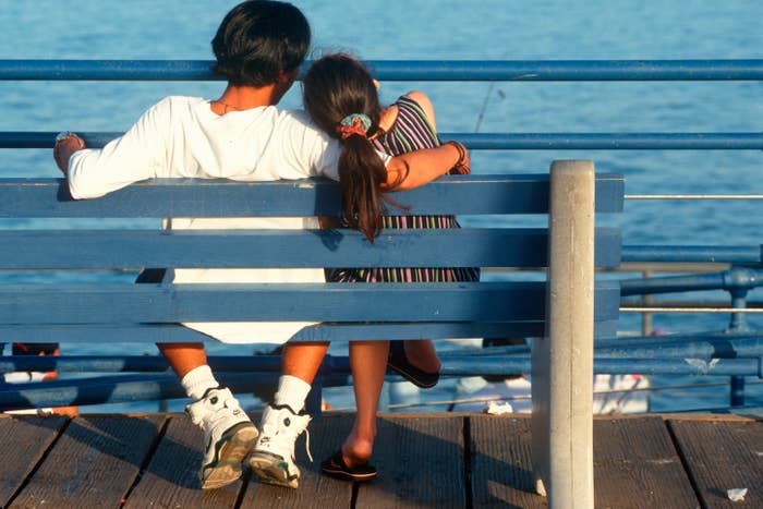 Two people sit closely on a bench, overlooking the water, suggesting a sense of companionship and affection