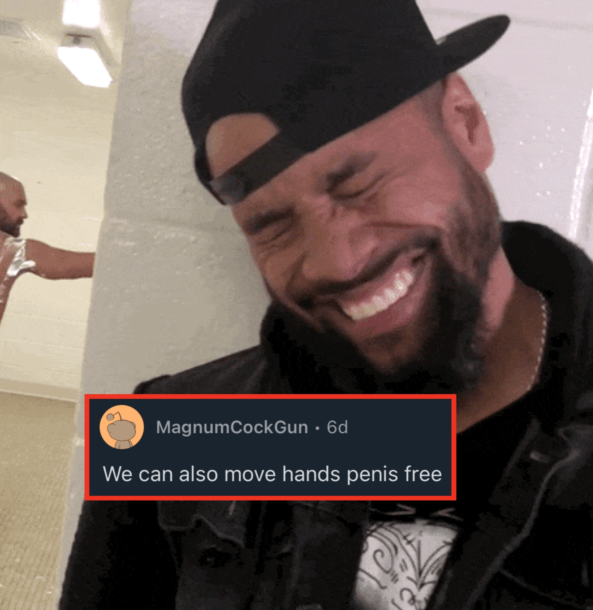 Man in a black cap and jacket smiles widely, leaning against a wall. Another person is visible in the background