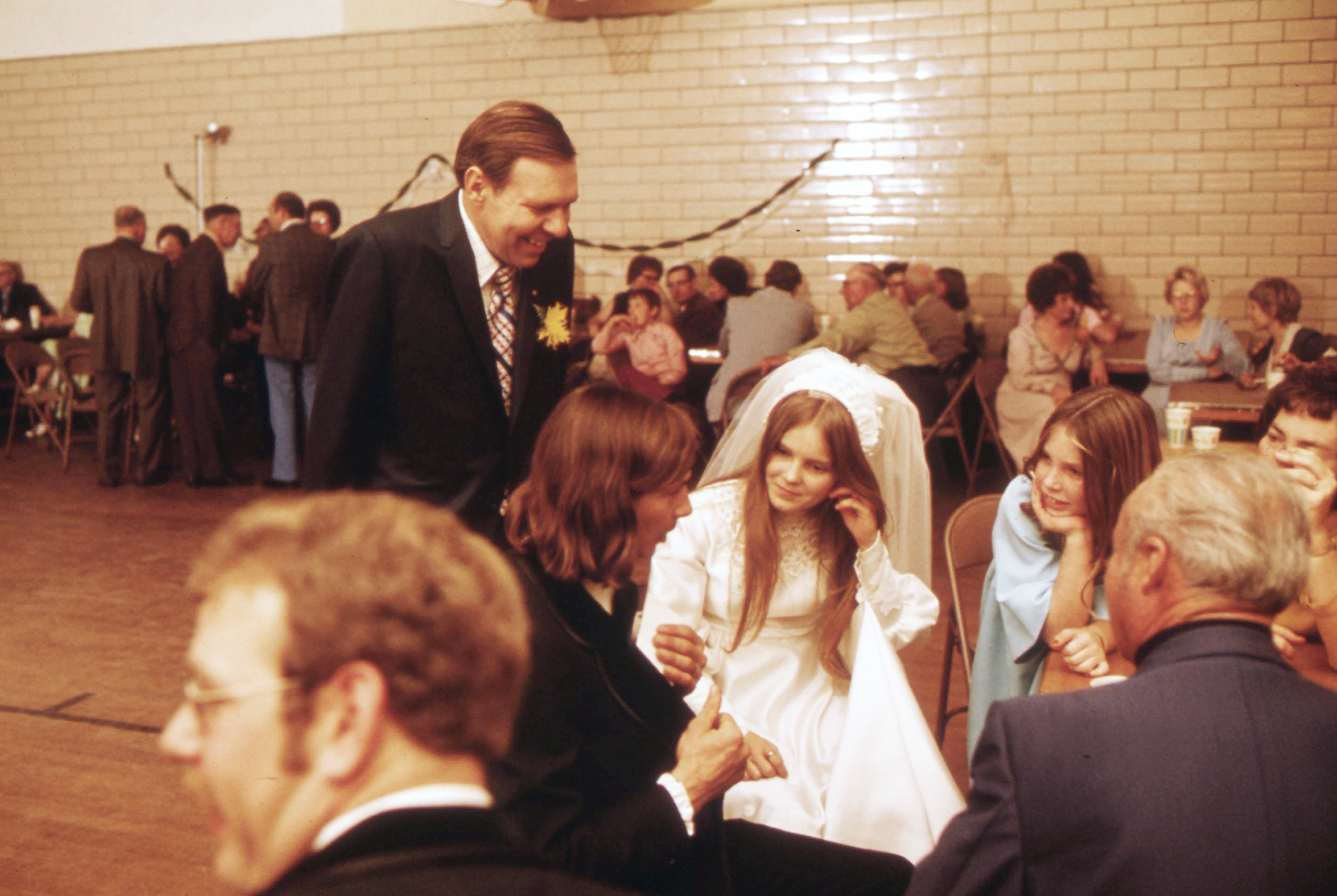 Group of people at a wedding reception, including a bride in a white dress and veil, engaged in conversation. Background shows seated guests