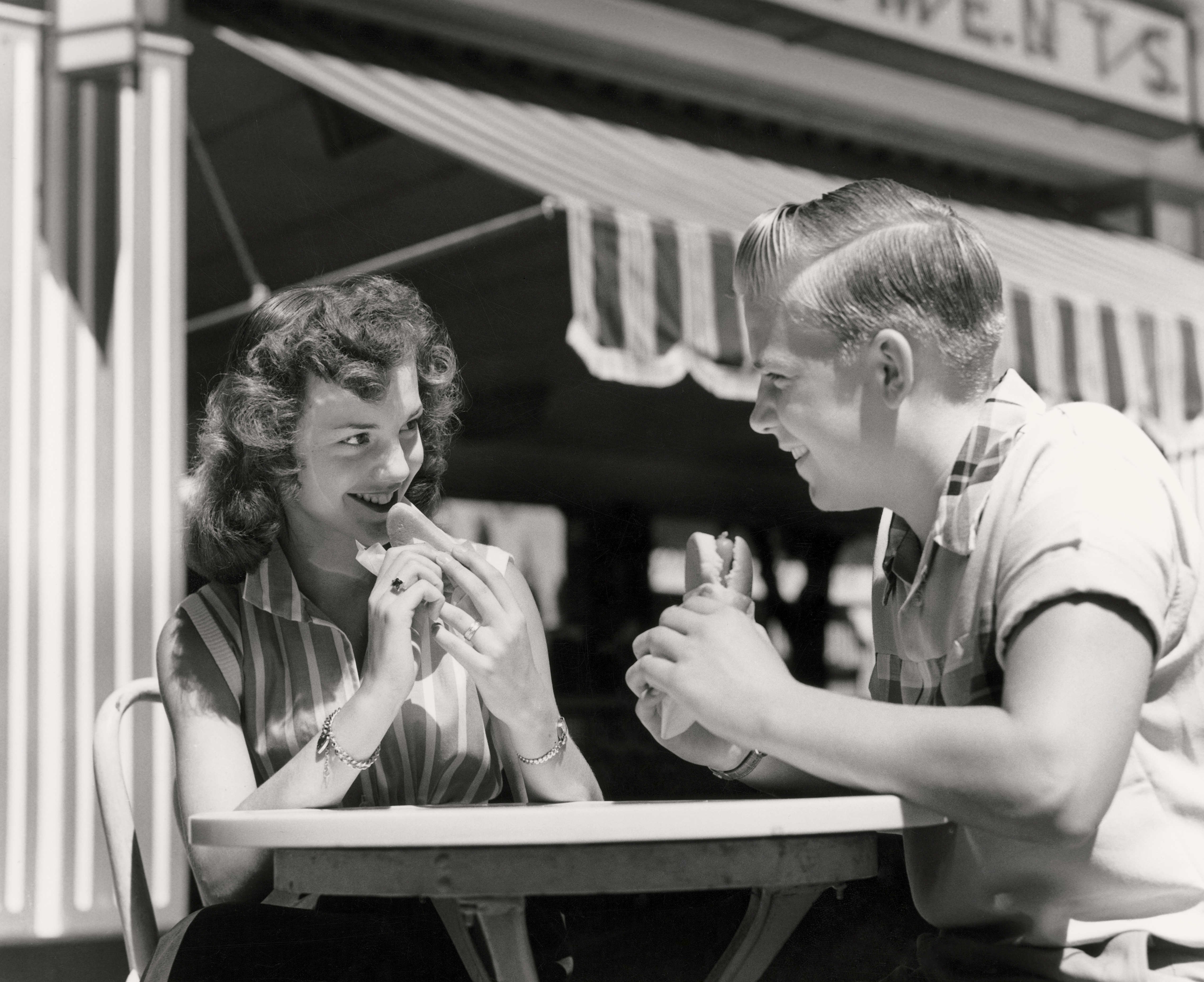 A vintage couple smiles and enjoys sharing hot dogs at an outdoor diner, evoking a playful, romantic vibe