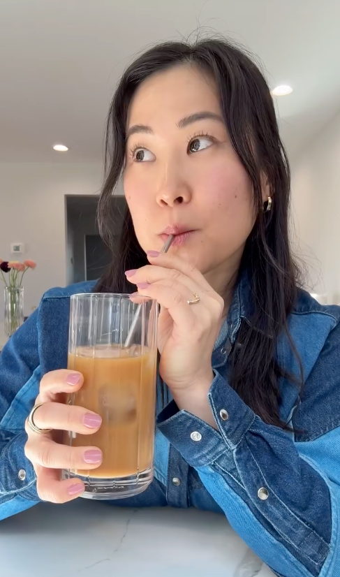 Person in denim shirt sipping iced coffee through a straw. Looking thoughtfully upward. Flowers visible in the background
