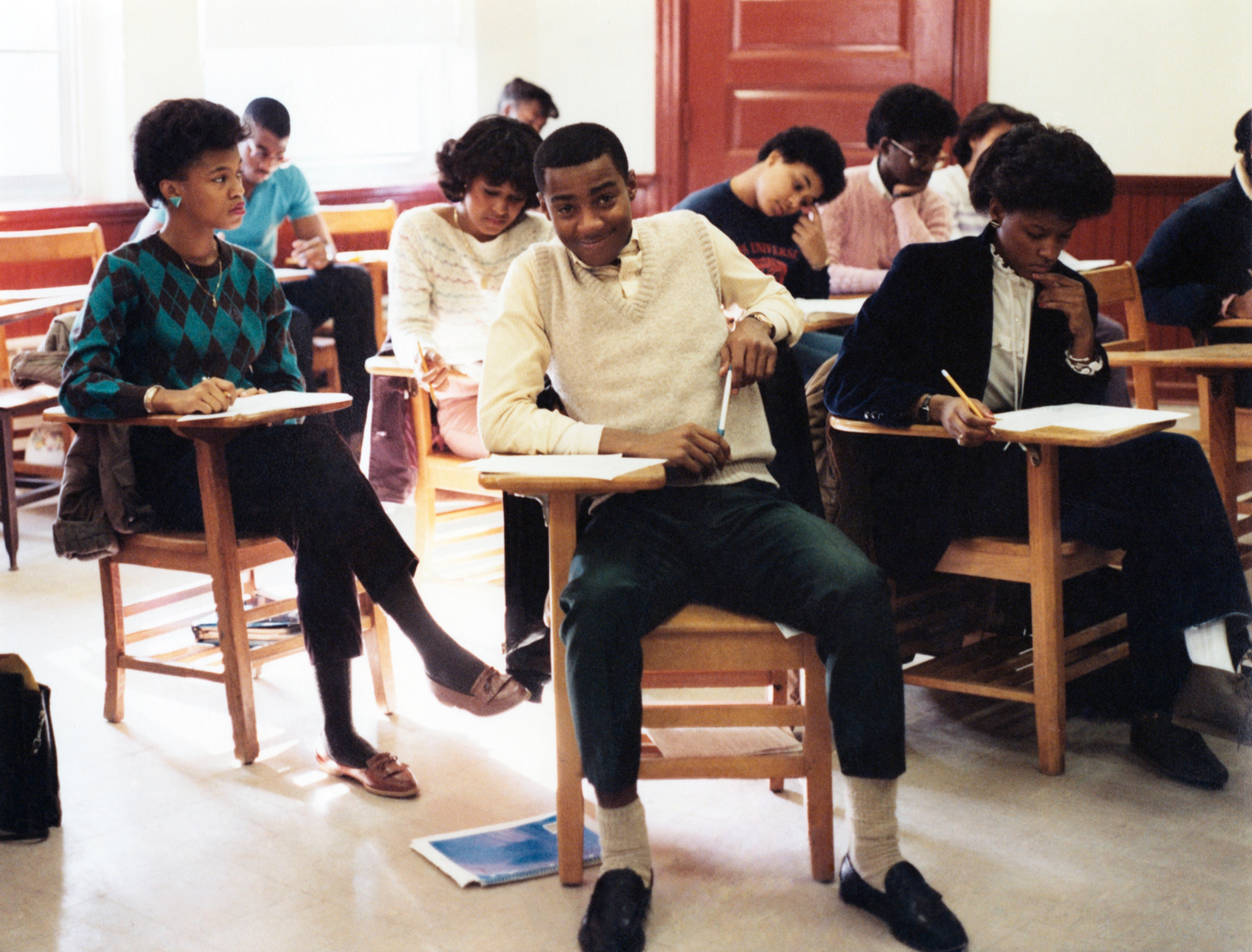 Students sit in a classroom at wooden desks, engaging with their work. The atmosphere appears focused and studious