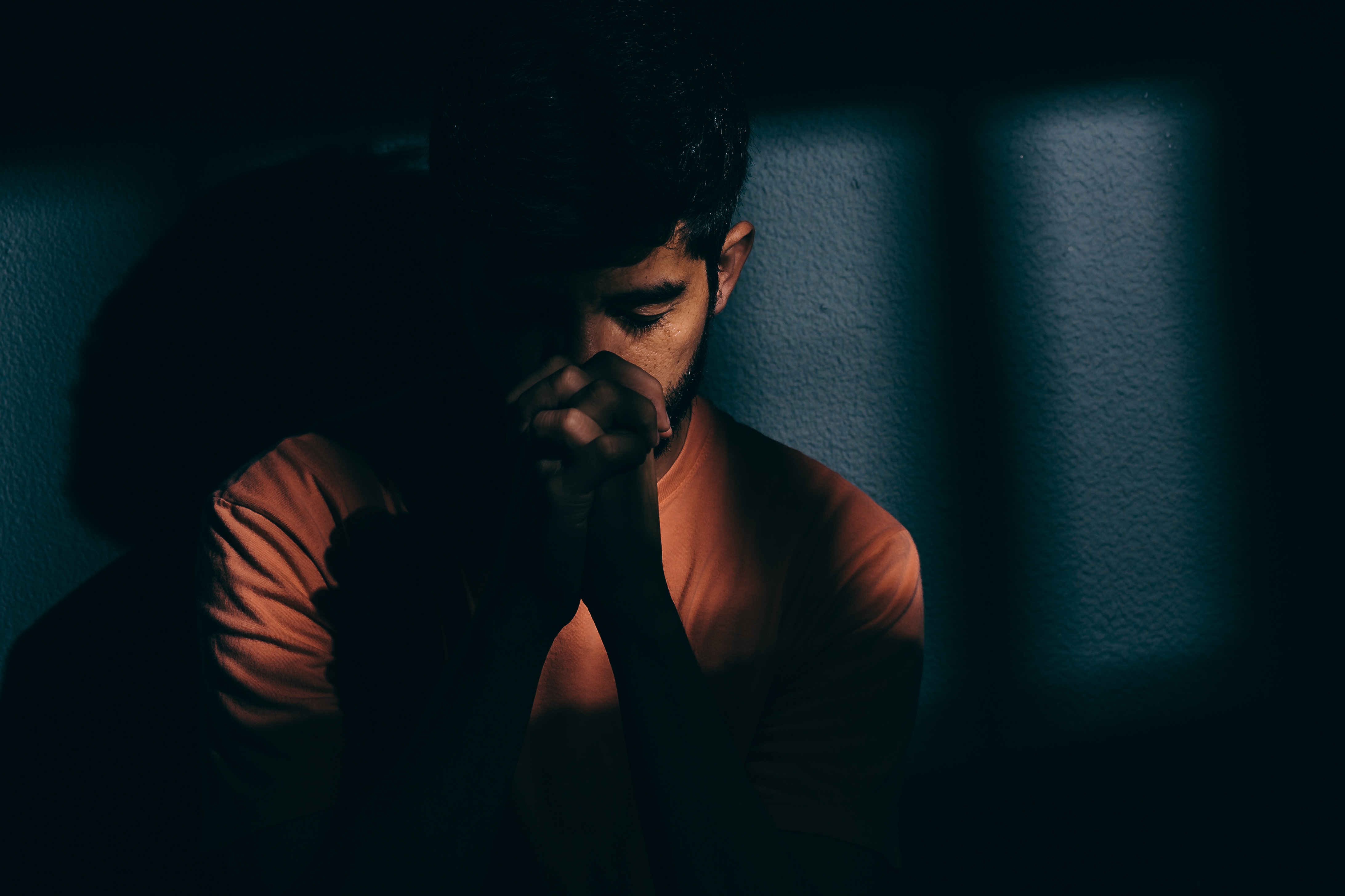 A person sits in deep thought, hands clasped, in a shadowy setting, conveying a reflective mood