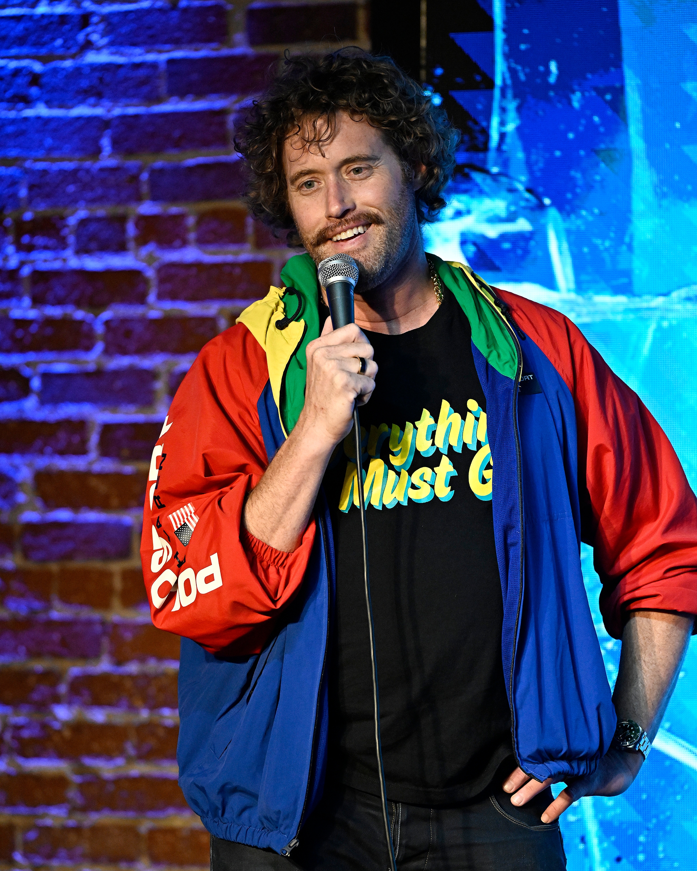 Person on stage performing stand-up, wearing a vibrant jacket and holding a microphone, in front of a brick wall with a blue, lit backdrop