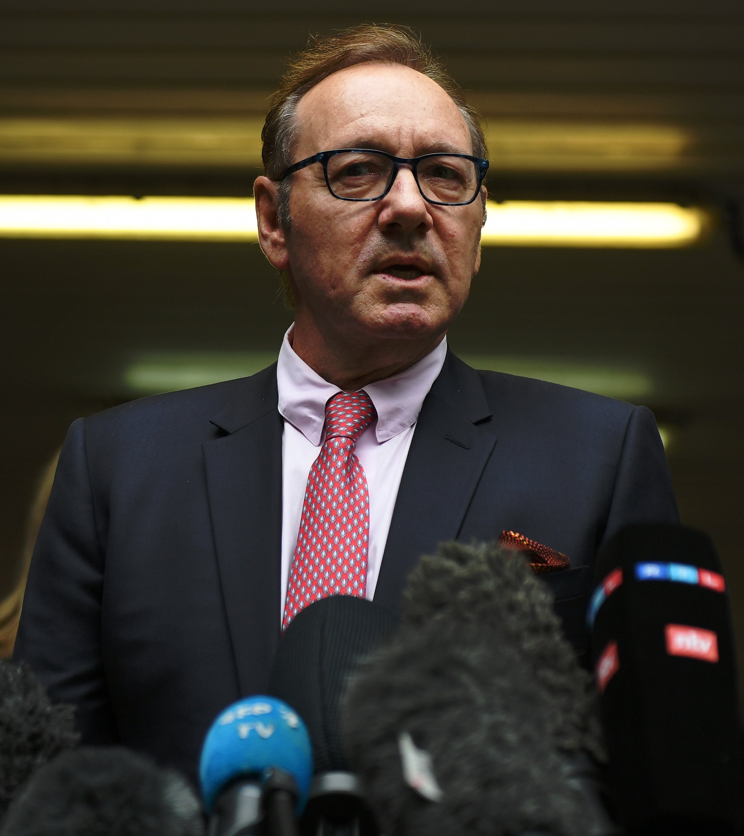 Person in a suit and tie stands in front of microphones, appearing to address a press conference