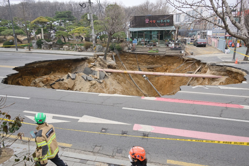 A large sinkhole has appeared on a city street, with emergency workers assessing the area. Nearby buildings and traffic are unaffected