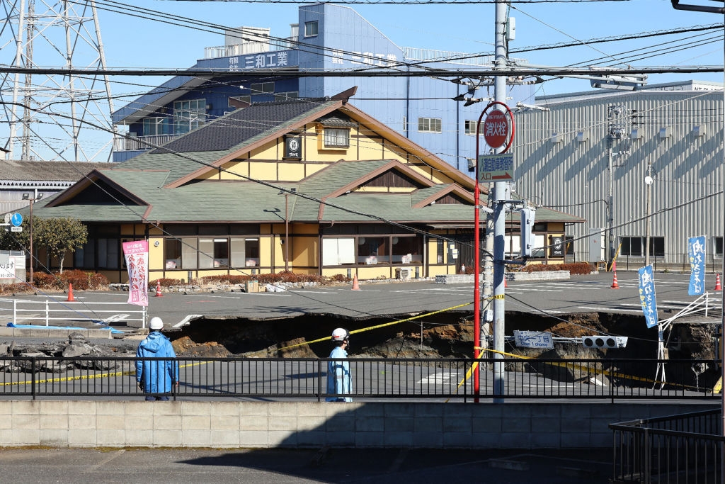 Workers in blue uniforms assess a road collapse in front of a traditional-style building, marked off with caution tape
