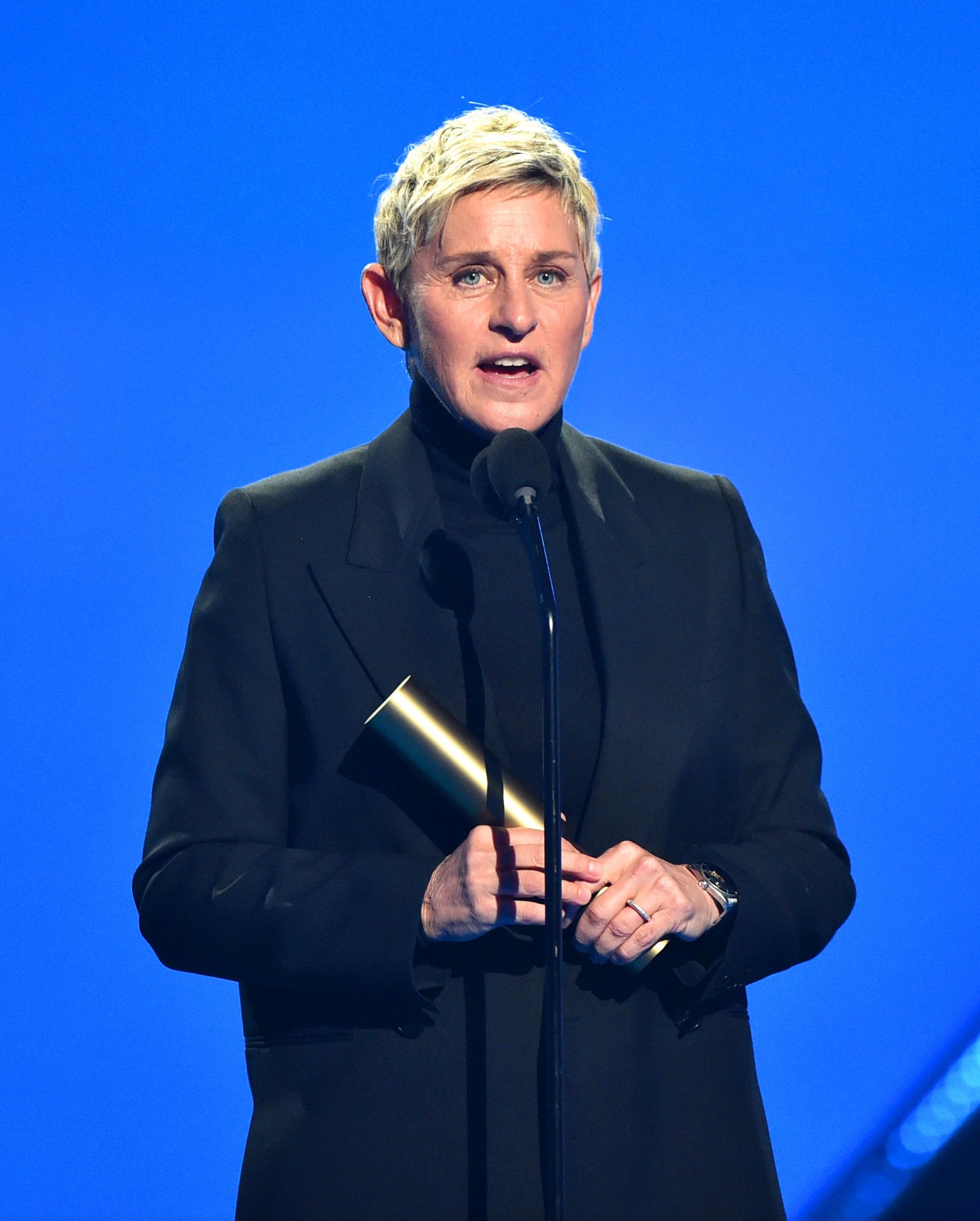 Person in a dark suit holding an award, speaking on stage at an event