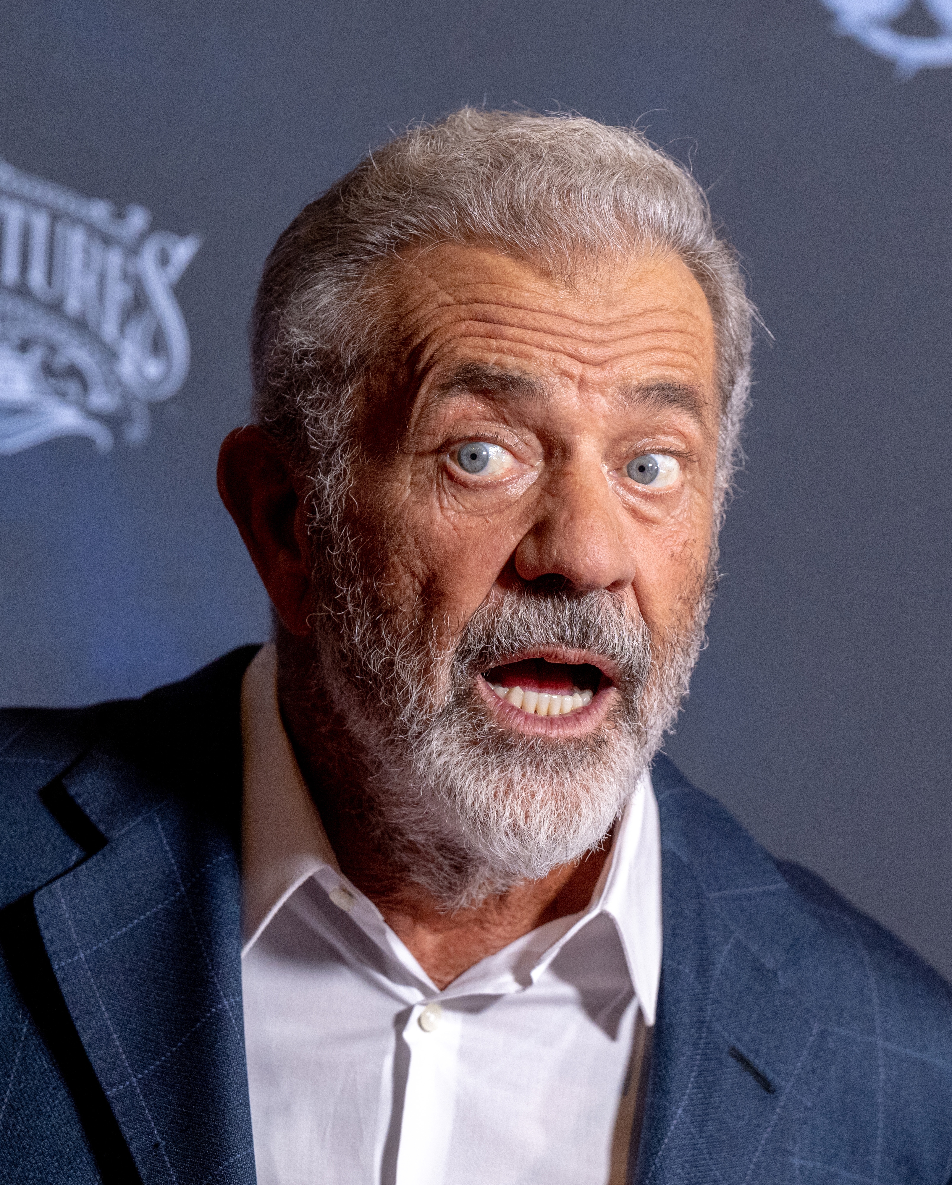A man with a beard looks surprised on a red carpet, wearing a suit jacket over a white shirt